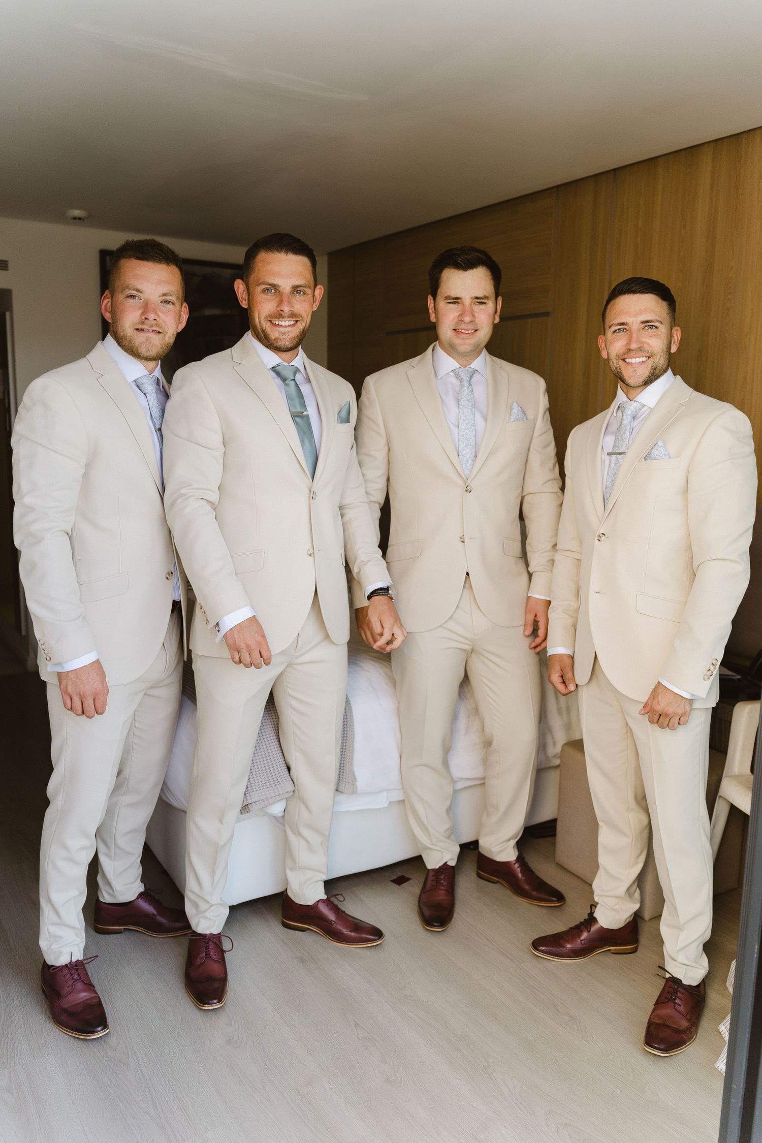 Four groomsmen in coordinating cream suits and blue ties stand together in a hallway.
