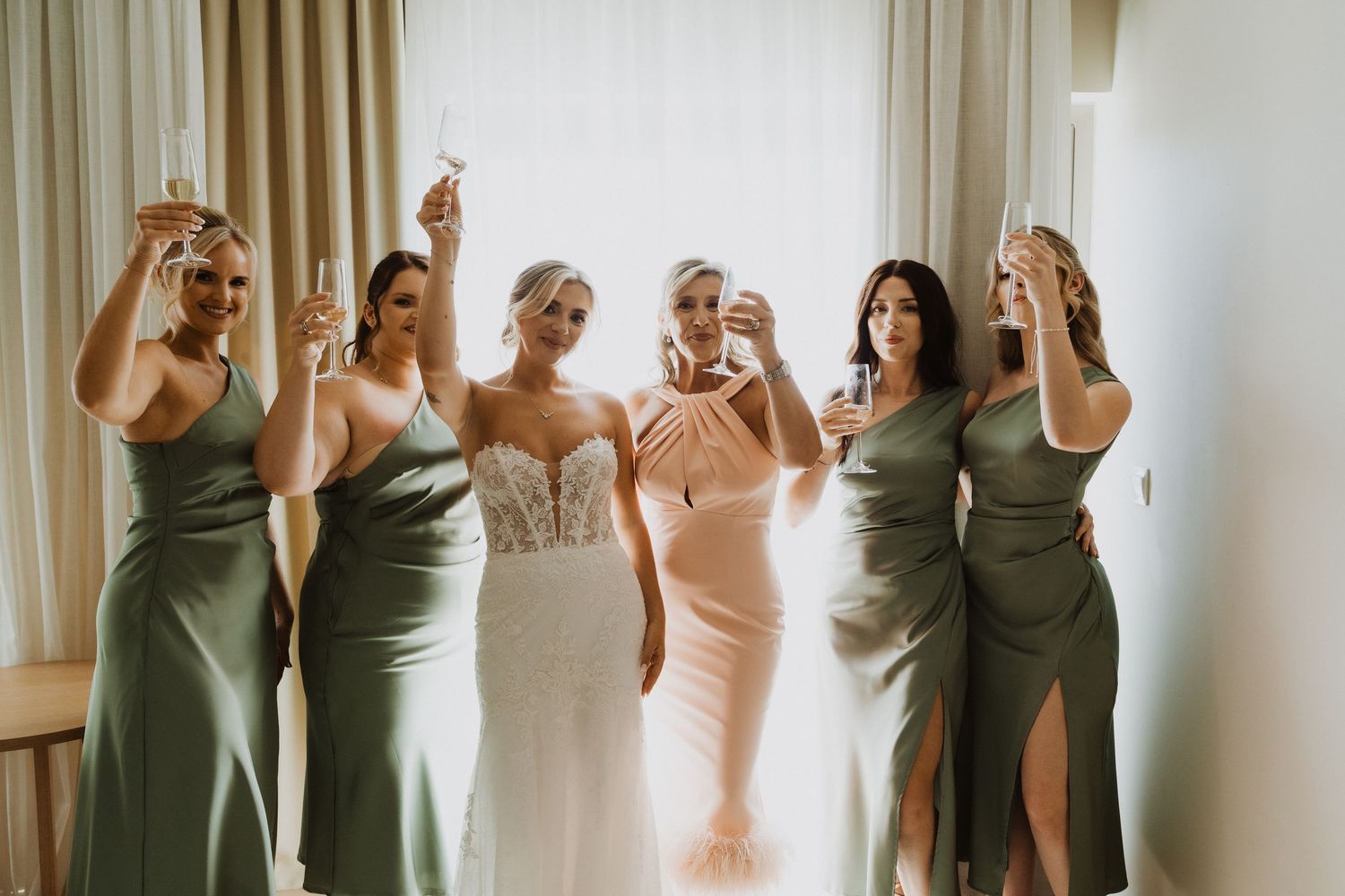 Wedding party members in coordinating sage and blush dresses pose against a white wall.