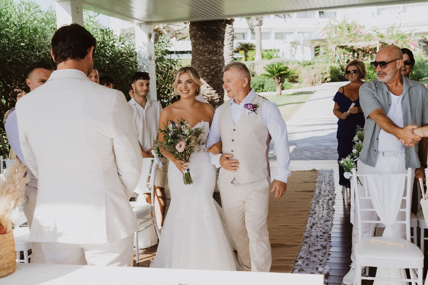 Wedding party members proceed down a decorated aisle at an outdoor covered venue.