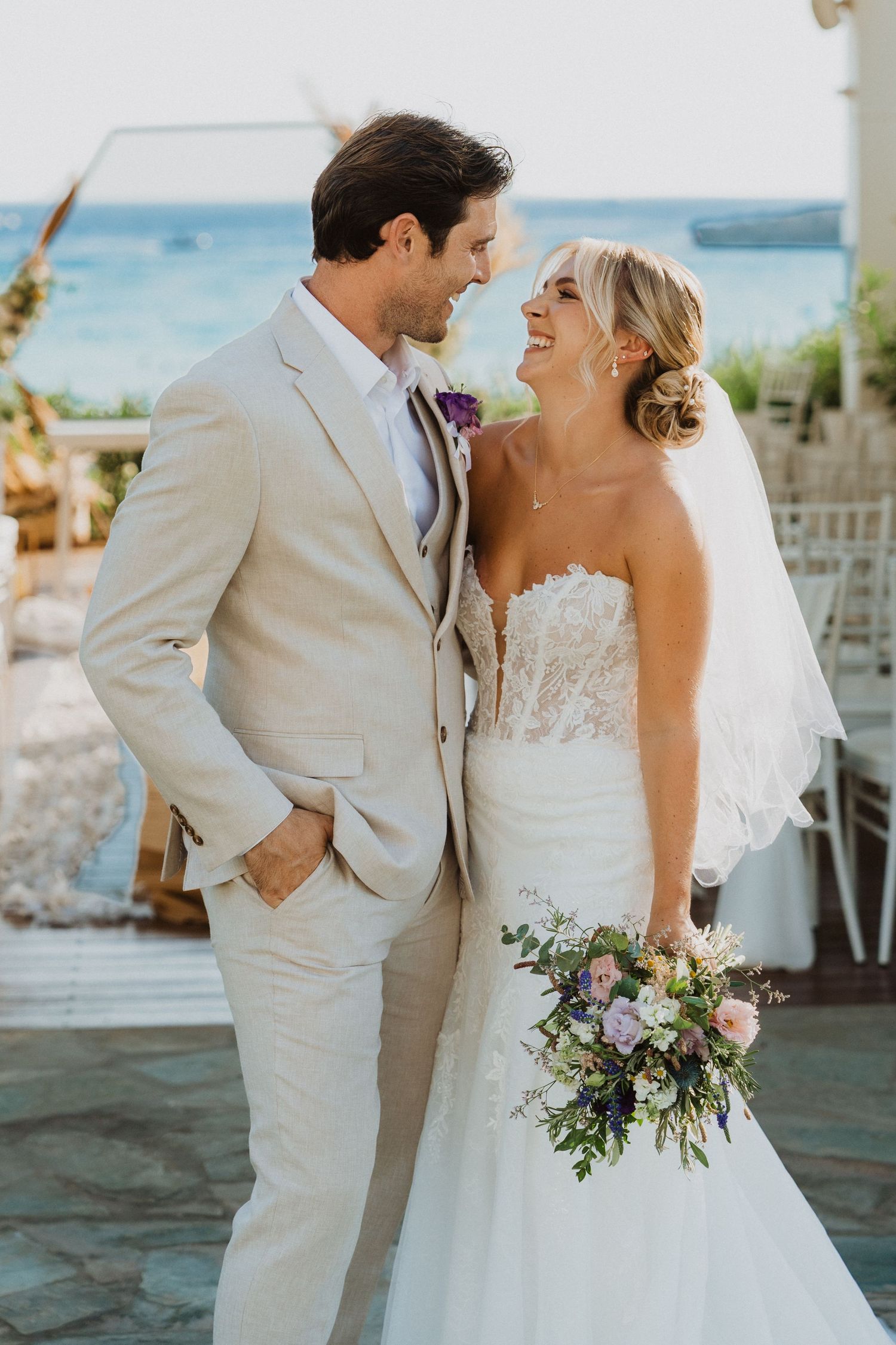 A bride in a white lace gown and groom in light suit embrace during their tropical beach wedding.