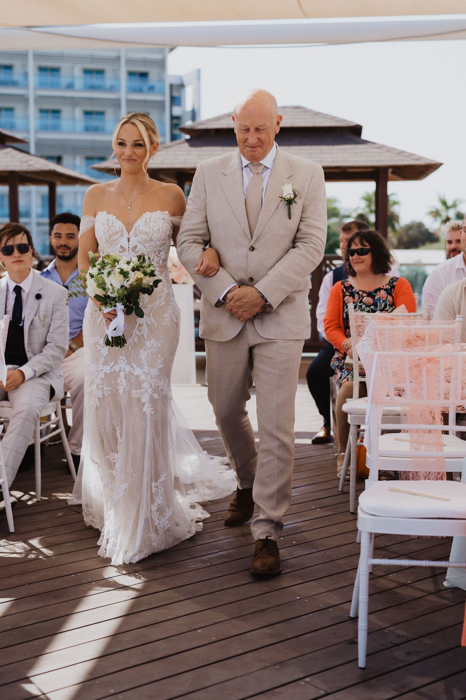 A wedding ceremony takes place on a wooden deck with white chairs and floral decorations by the ocean.