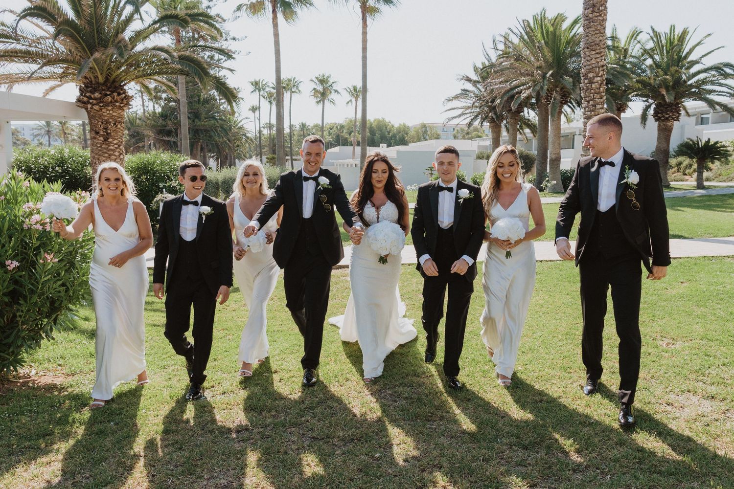 Wedding party members jumping joyfully on grass with palm trees in background.