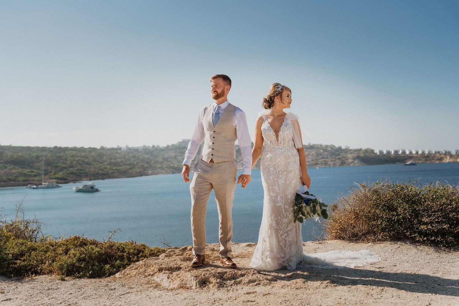Couple walks along scenic coastal cliffs in light colored wedding attire with ocean views in background.