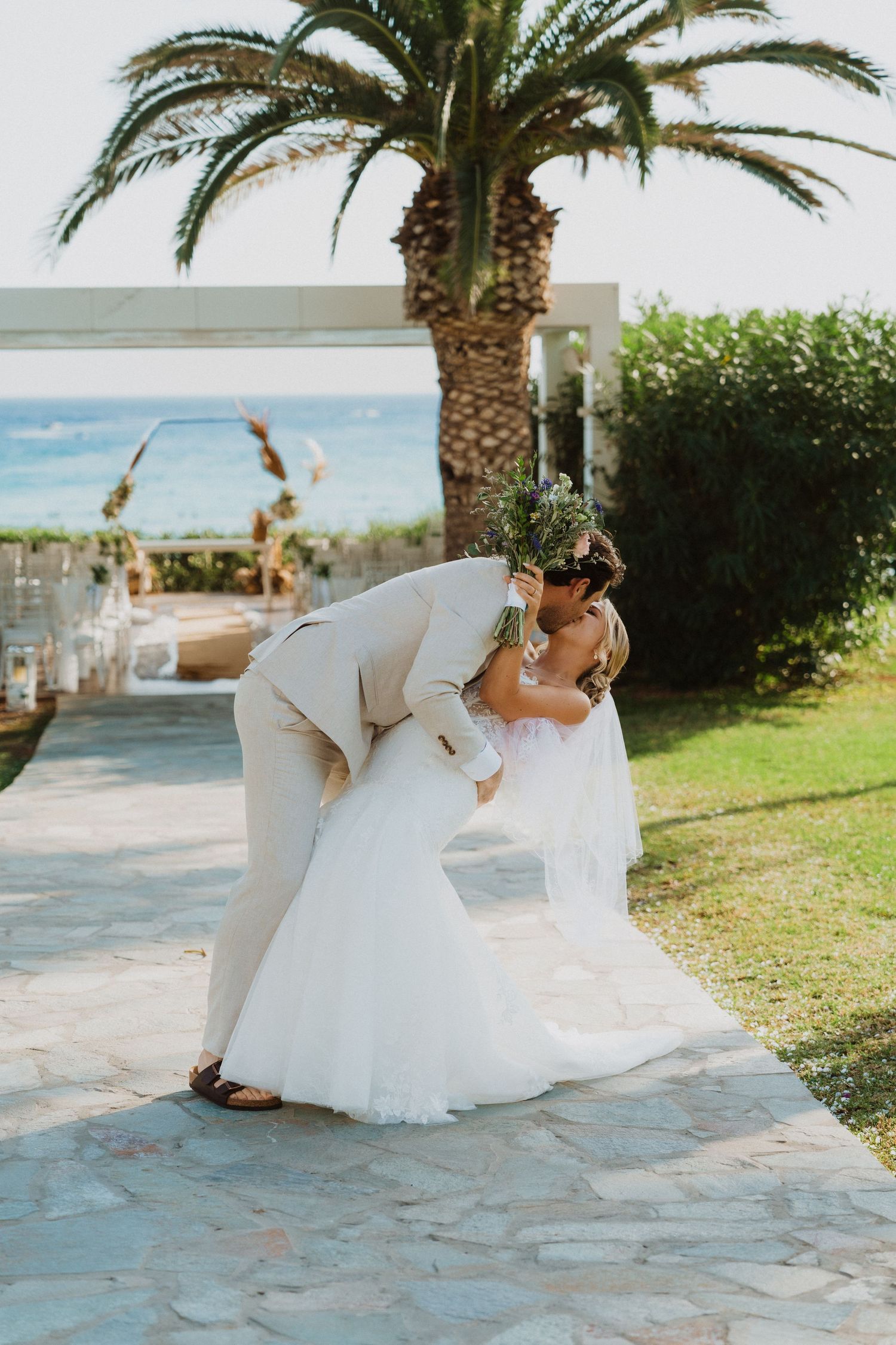 Wedding couple dips and poses playfully near palm trees on stone pathway.