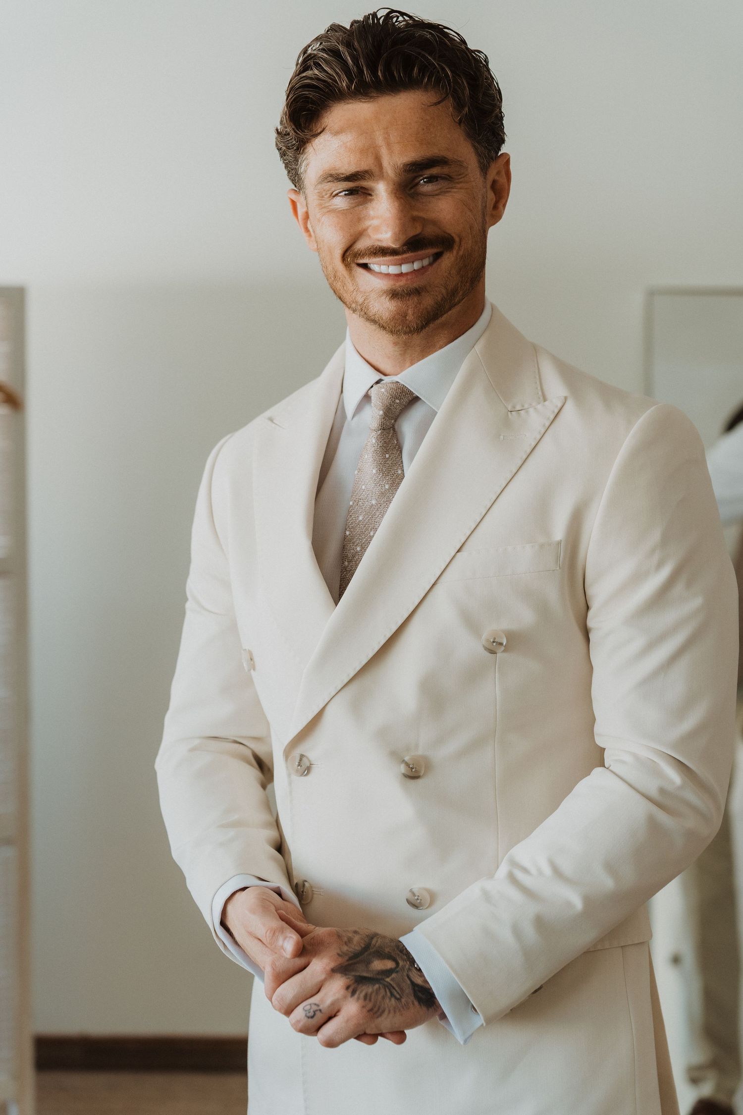 A person in a crisp white double-breasted suit and tie stands confidently in a bright interior setting.