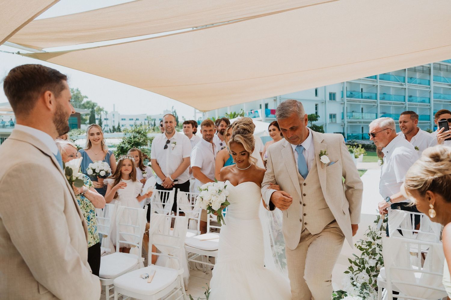 Bride walks down the aisle with her father at an outdoor beach wedding ceremony with guests in summer attire.