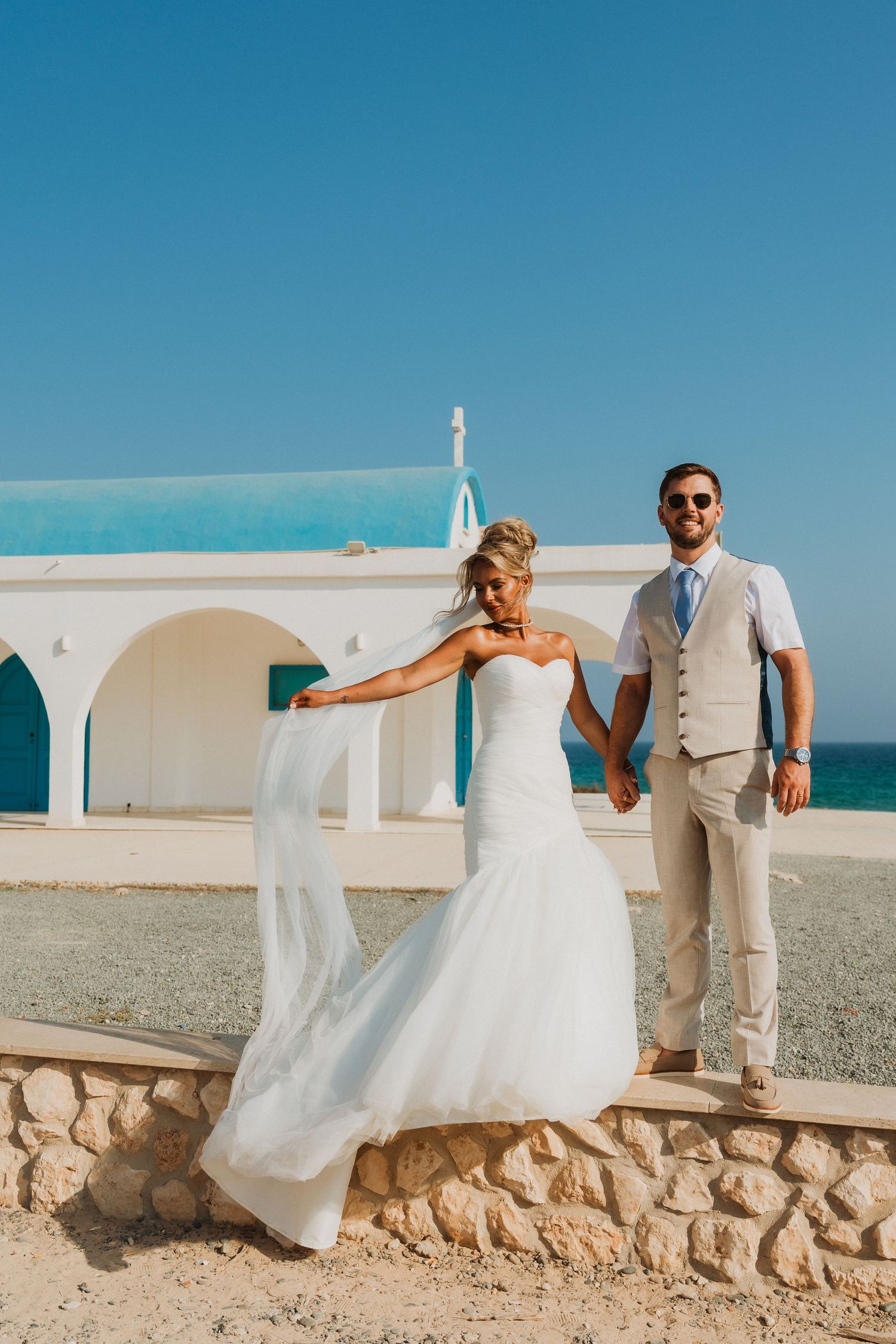 Bride and groom holding hands at a seaside wedding venue with a white and blue chapel in the background.