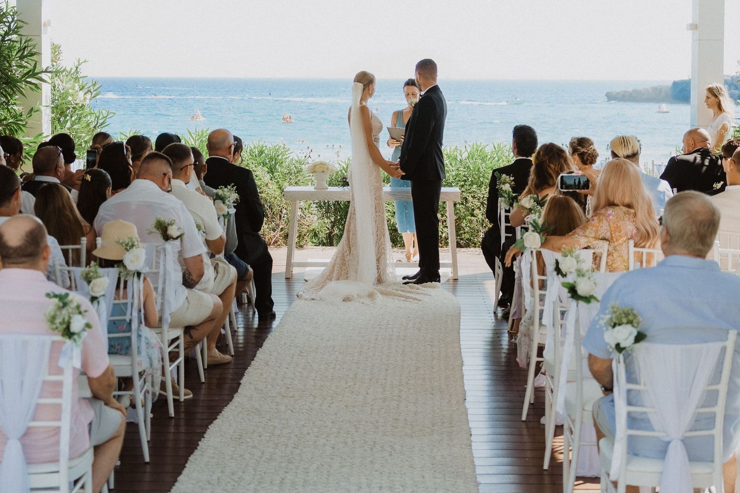 Wedding ceremony setup on a beach deck with white chairs and ocean views in the background.