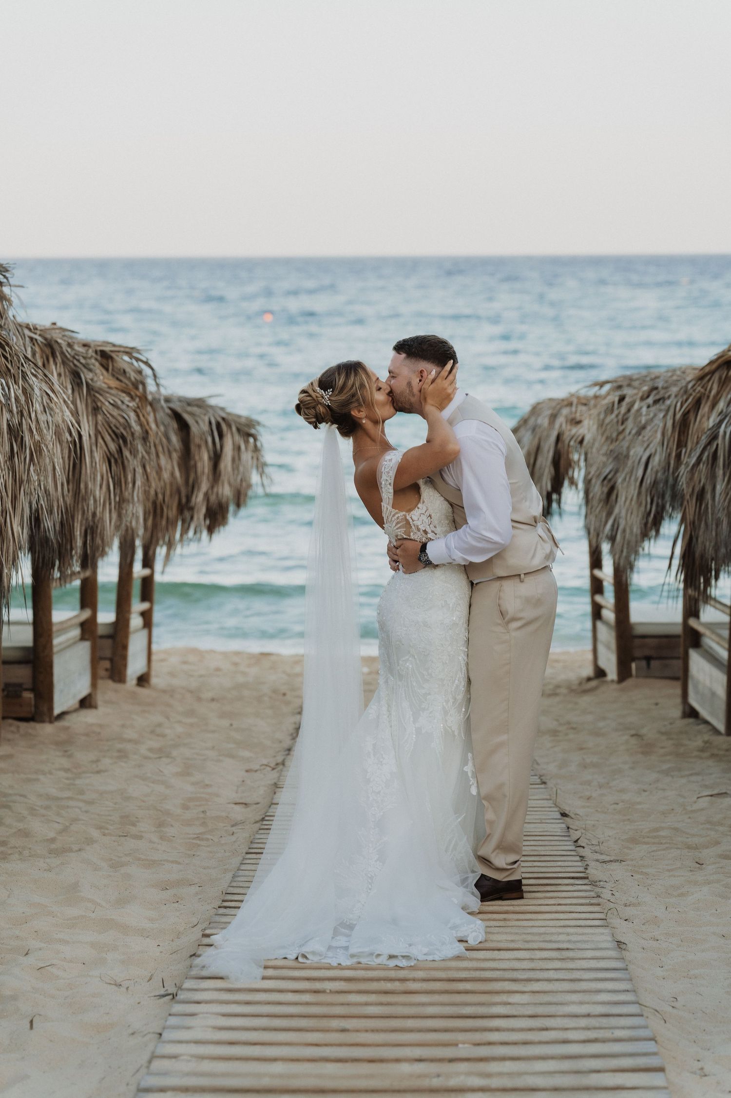 A couple in wedding attire shares an intimate moment on a beach boardwalk lined with thatched umbrellas near the ocean.