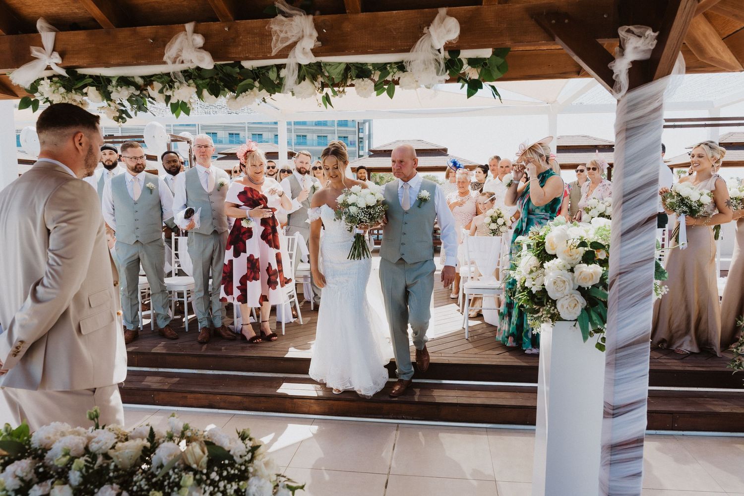 A joyful celebration at an outdoor wedding ceremony with white floral decorations and greenery under a wooden pergola.