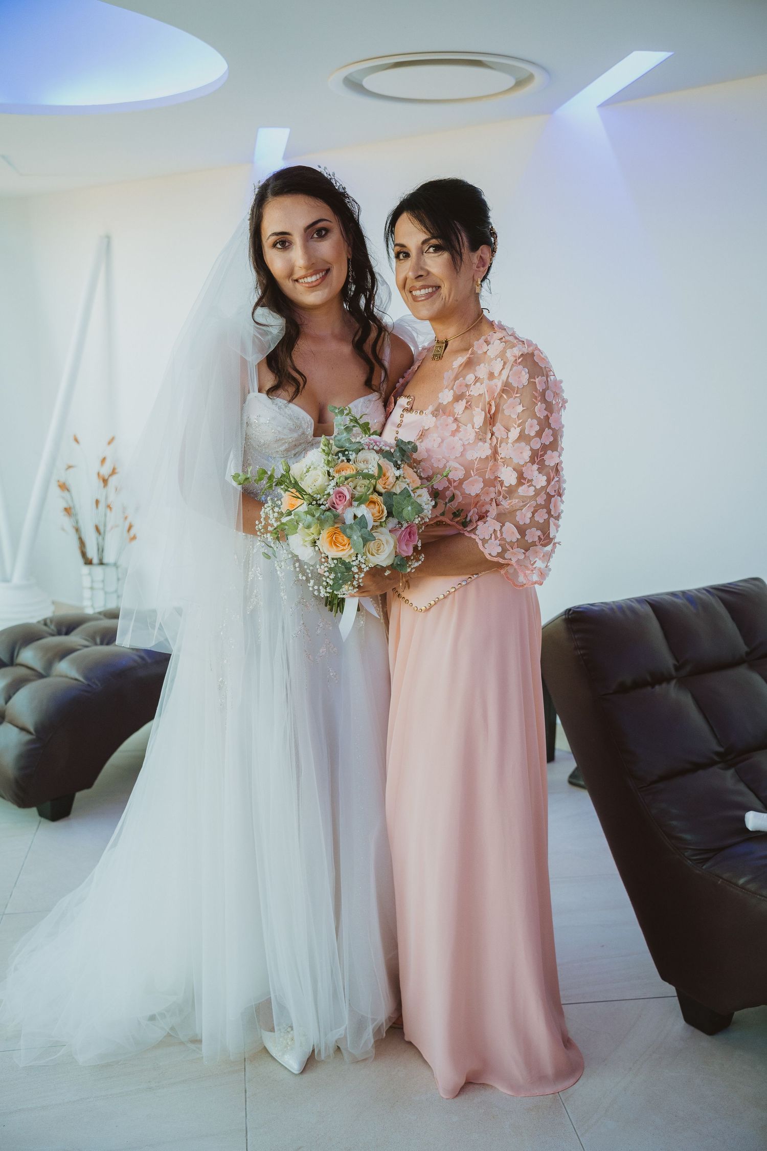 Bride in white gown poses with bridesmaid in pink floral dress holding pastel bouquet.