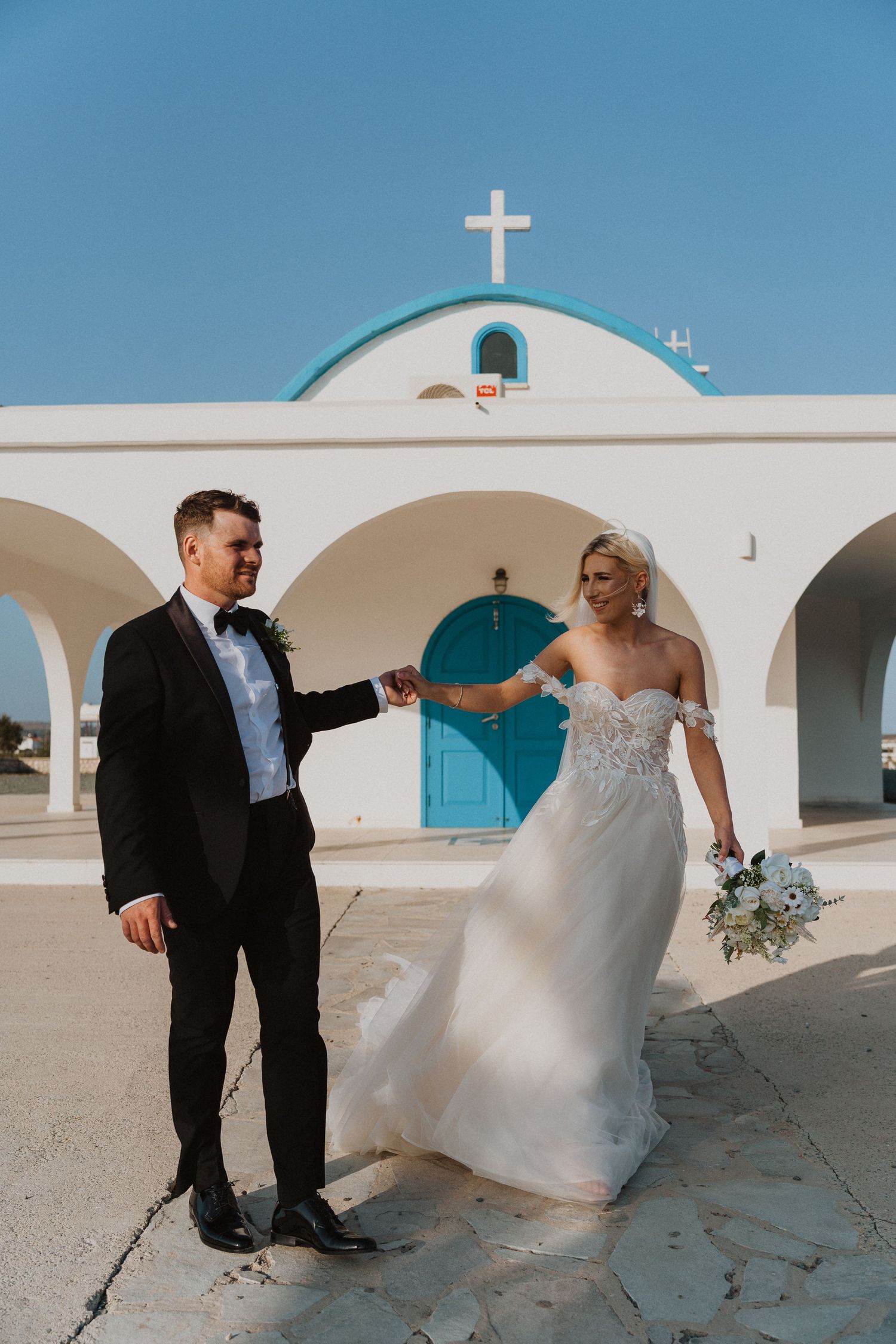 Newlyweds share playful moment outside white Mediterranean church with turquoise door and cross.