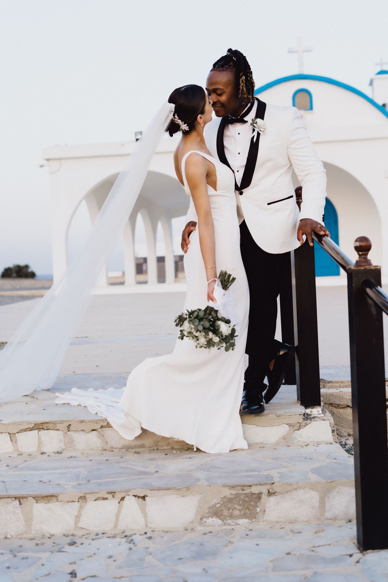 Newlyweds share an intimate moment on stone steps of a Greek Orthodox church with white walls.