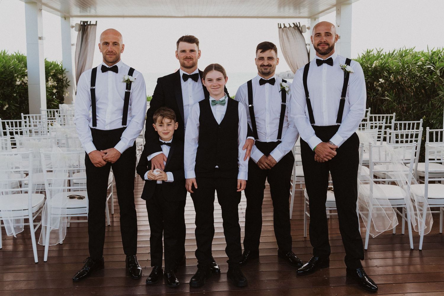 Groomsmen in black suits with suspenders pose in ceremony space with white chairs.