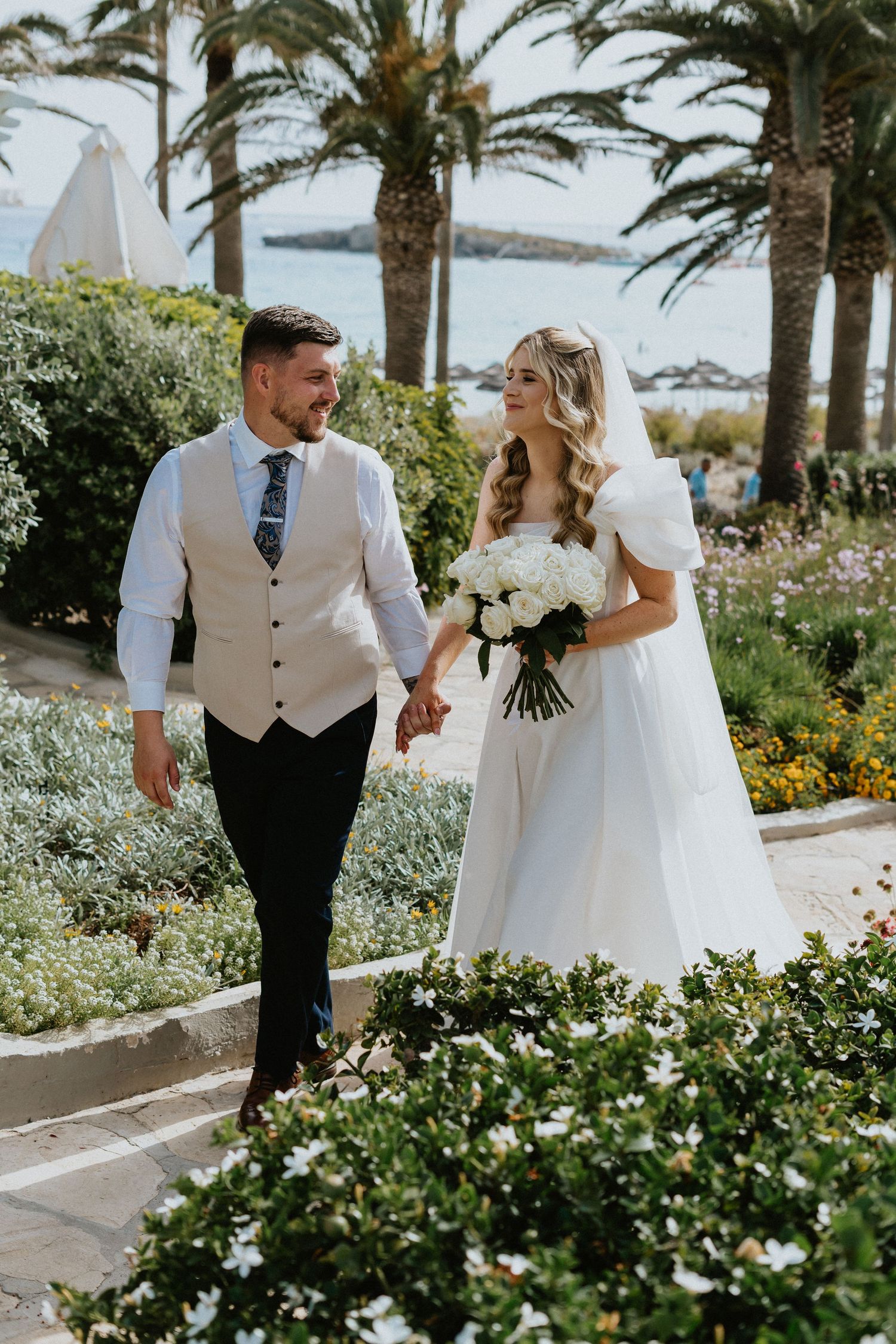 Couple walking through tropical garden pathway with palm trees and ocean backdrop.