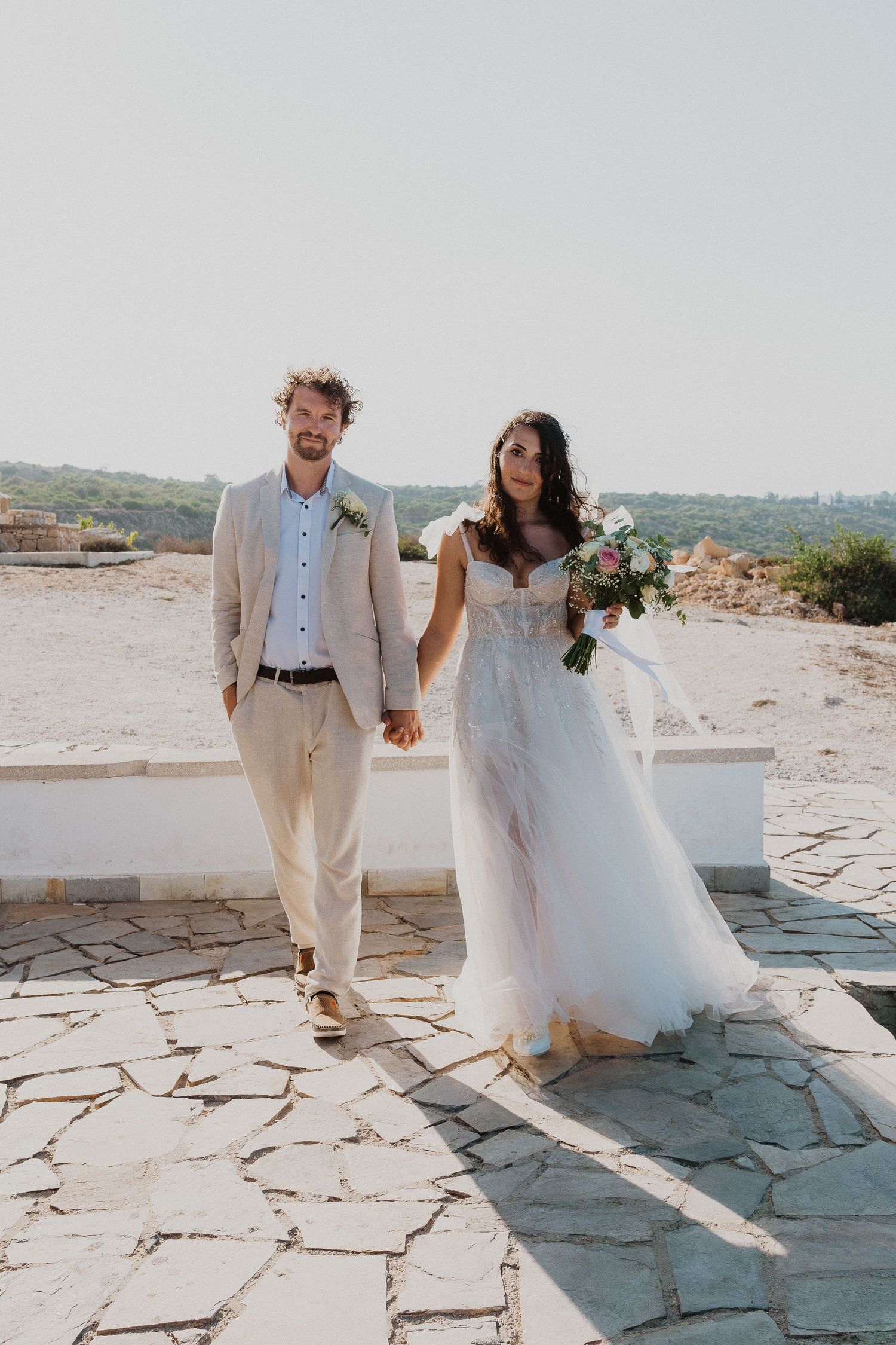 A newlywed couple walks together on a stone pathway overlooking the Mediterranean coast.