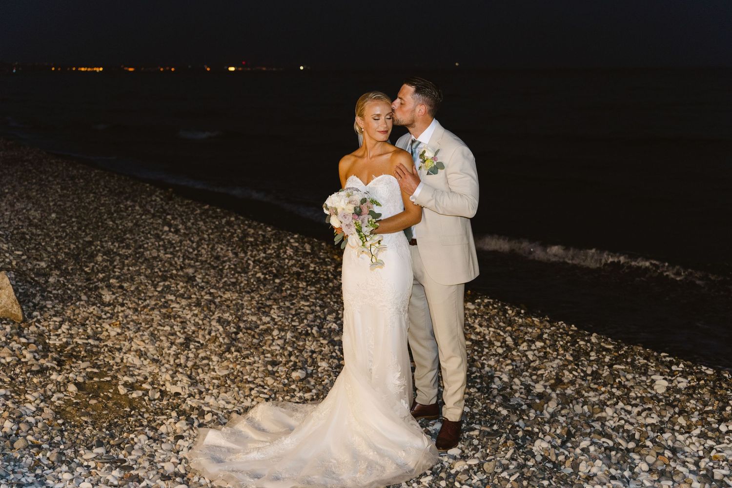 Beautiful beach wedding scene with couple embracing on pebbled shore at night.
