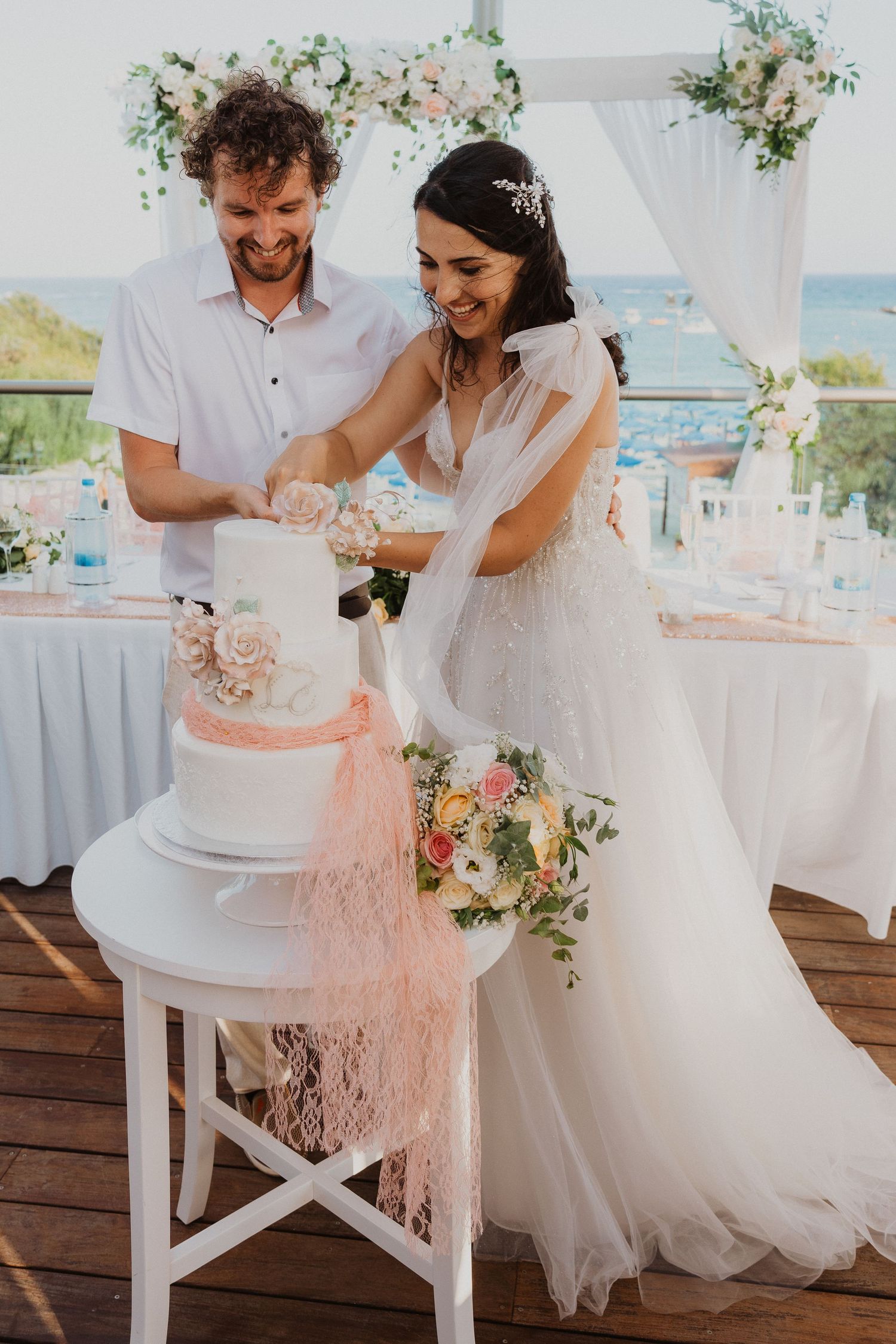 Newlyweds share a joyful moment while cutting their three-tiered wedding cake at a seaside venue.