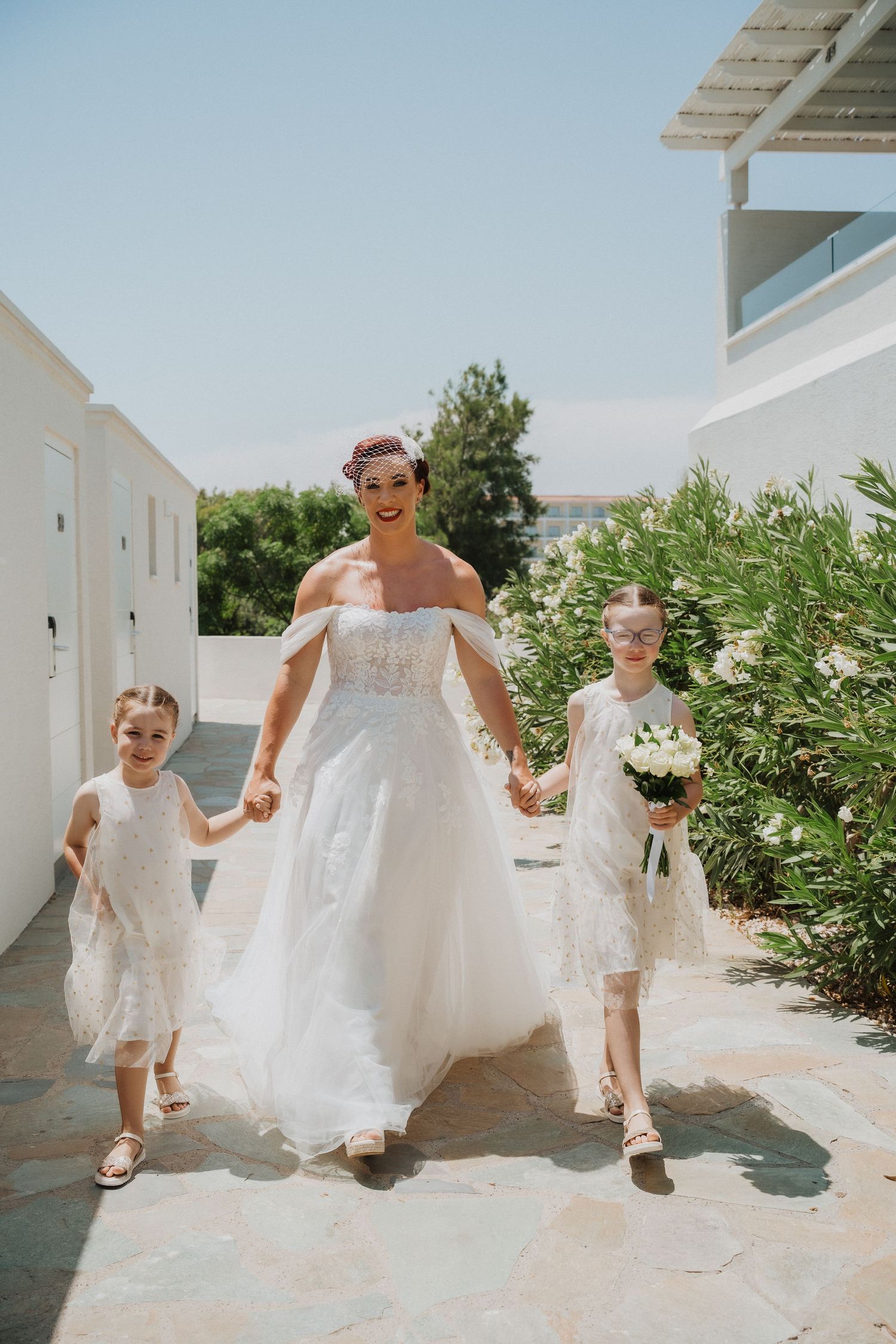 A wedding party walks along a sunny Mediterranean pathway with flowing white dresses in a garden setting.