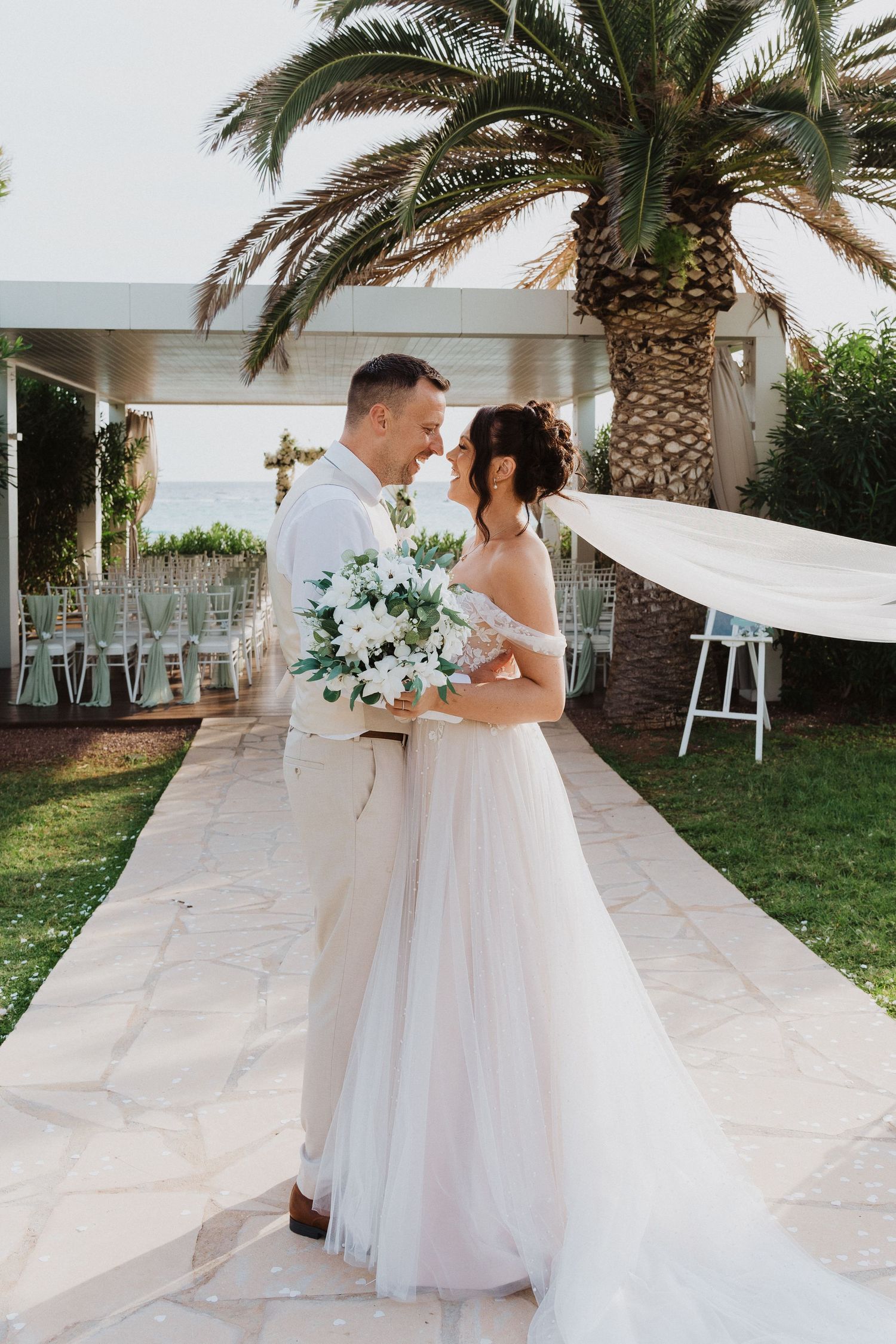 A romantic outdoor wedding ceremony with palm trees showcasing a couple in white attire with floral bouquet and flowing veil.