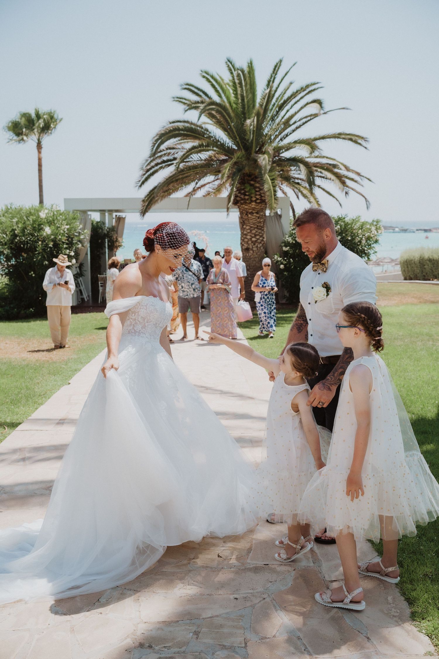 Elegant wedding scene with flowing white dress and young flower girls under palm trees.