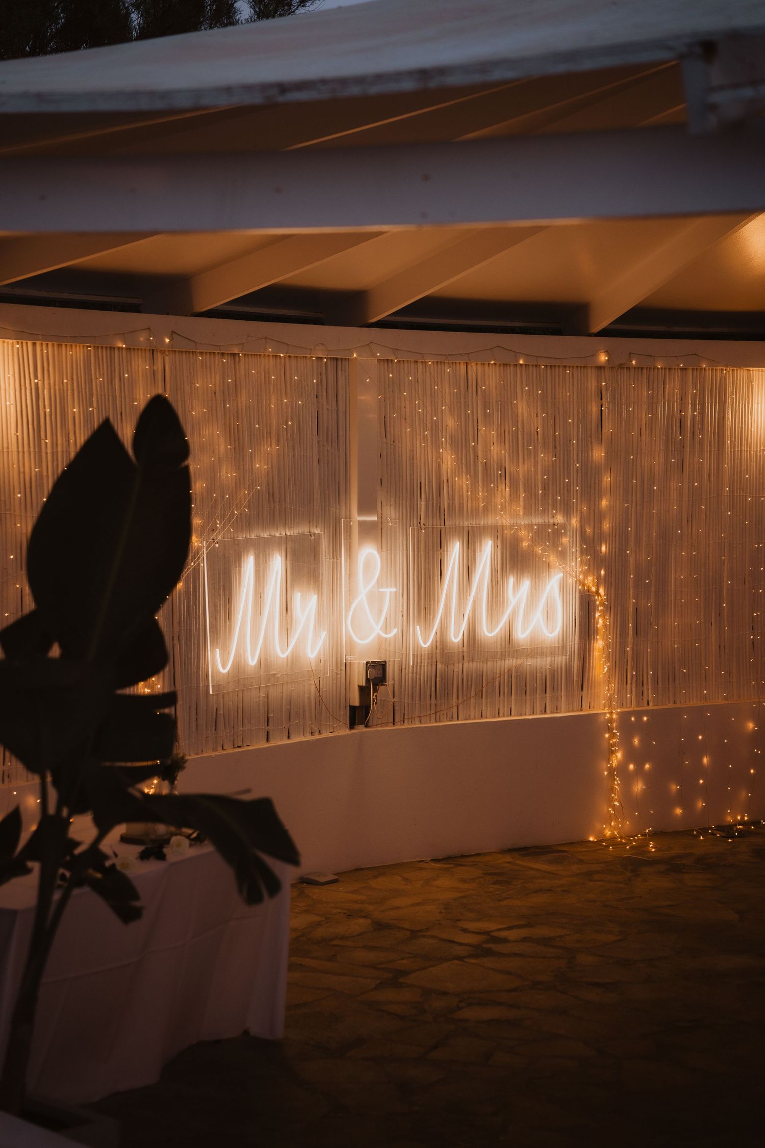 Illuminated 'Mr & Mrs' neon sign against fairy light backdrop at wedding reception.