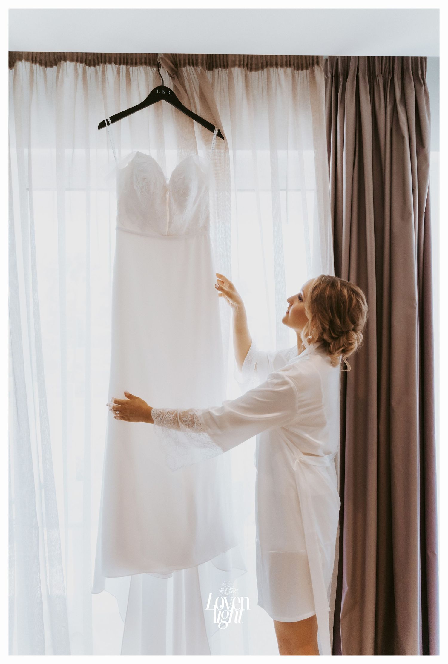 A white wedding dress hangs by a window while someone in a robe reaches up to touch it.
