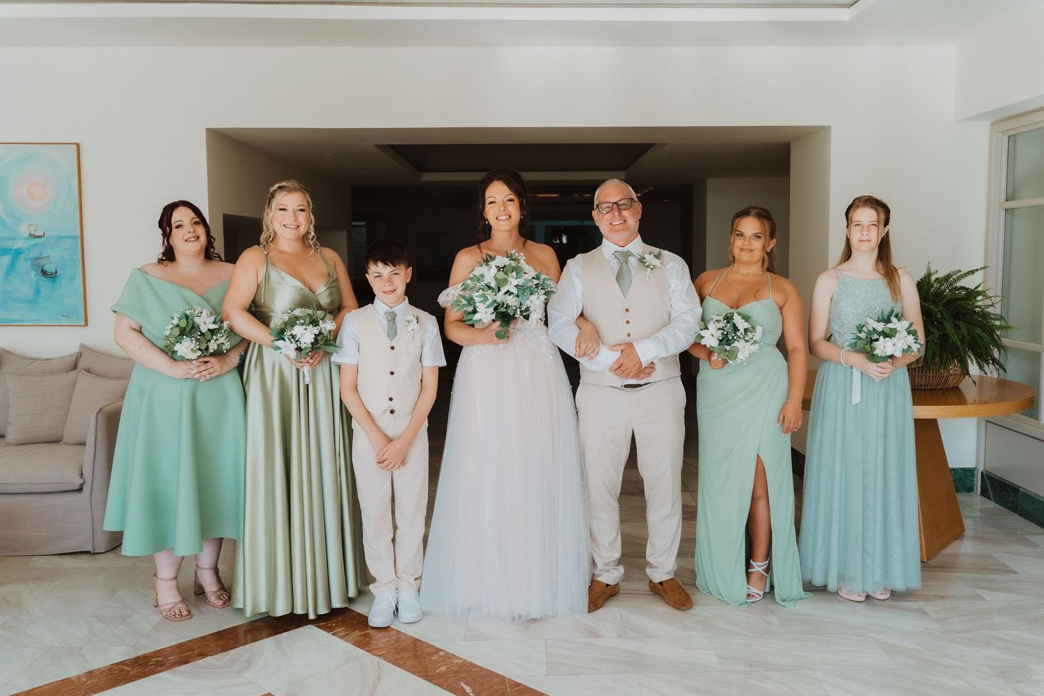 Wedding party poses together in mint green bridesmaid dresses and light colored suits in a bright interior space.