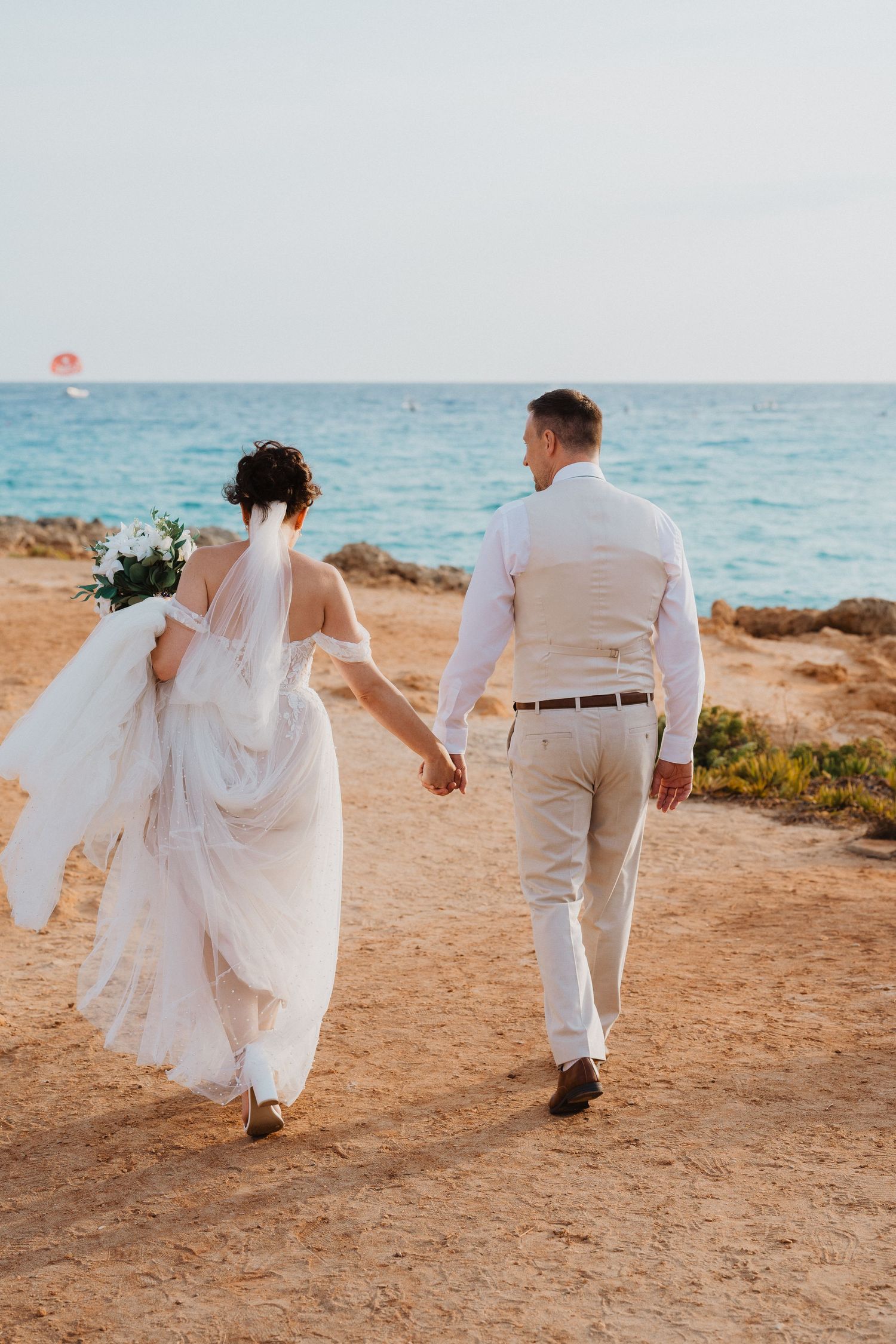 A newlywed couple walking hand in hand on a coastal cliff with the turquoise Mediterranean sea in the background.