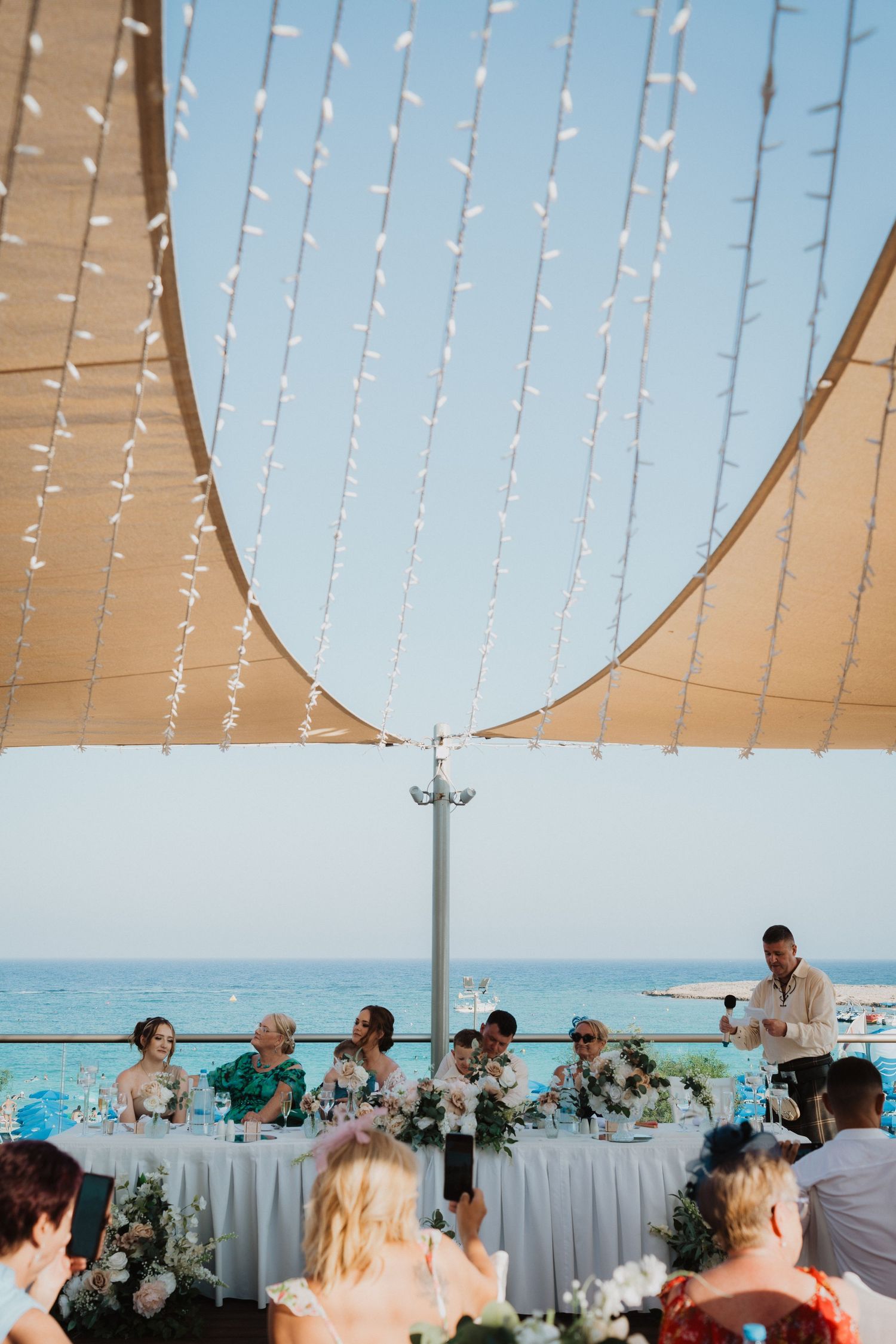 String lights drape over an oceanfront wedding reception as guests dine at decorated tables.
