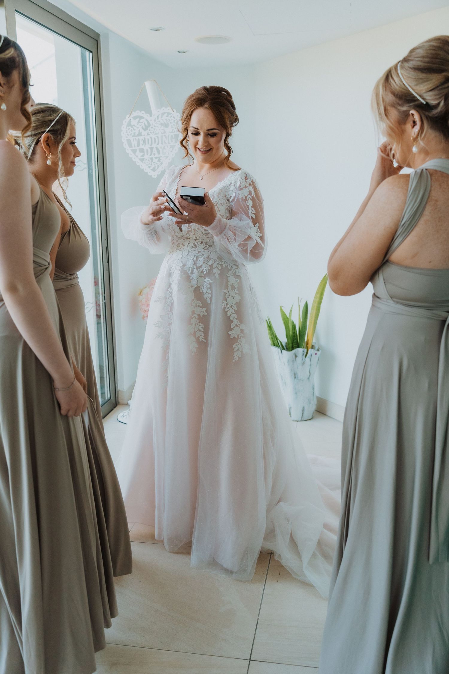 Elegant bride in full-length lace gown holds pink and white bouquet near window.