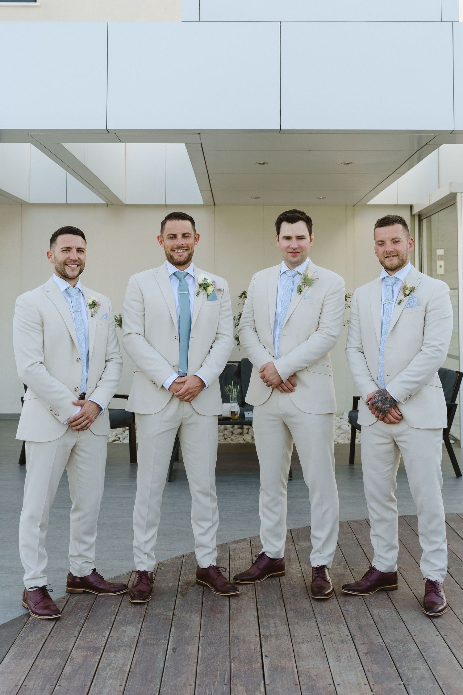 Four groomsmen in matching cream suits and blue ties pose on wooden deck.