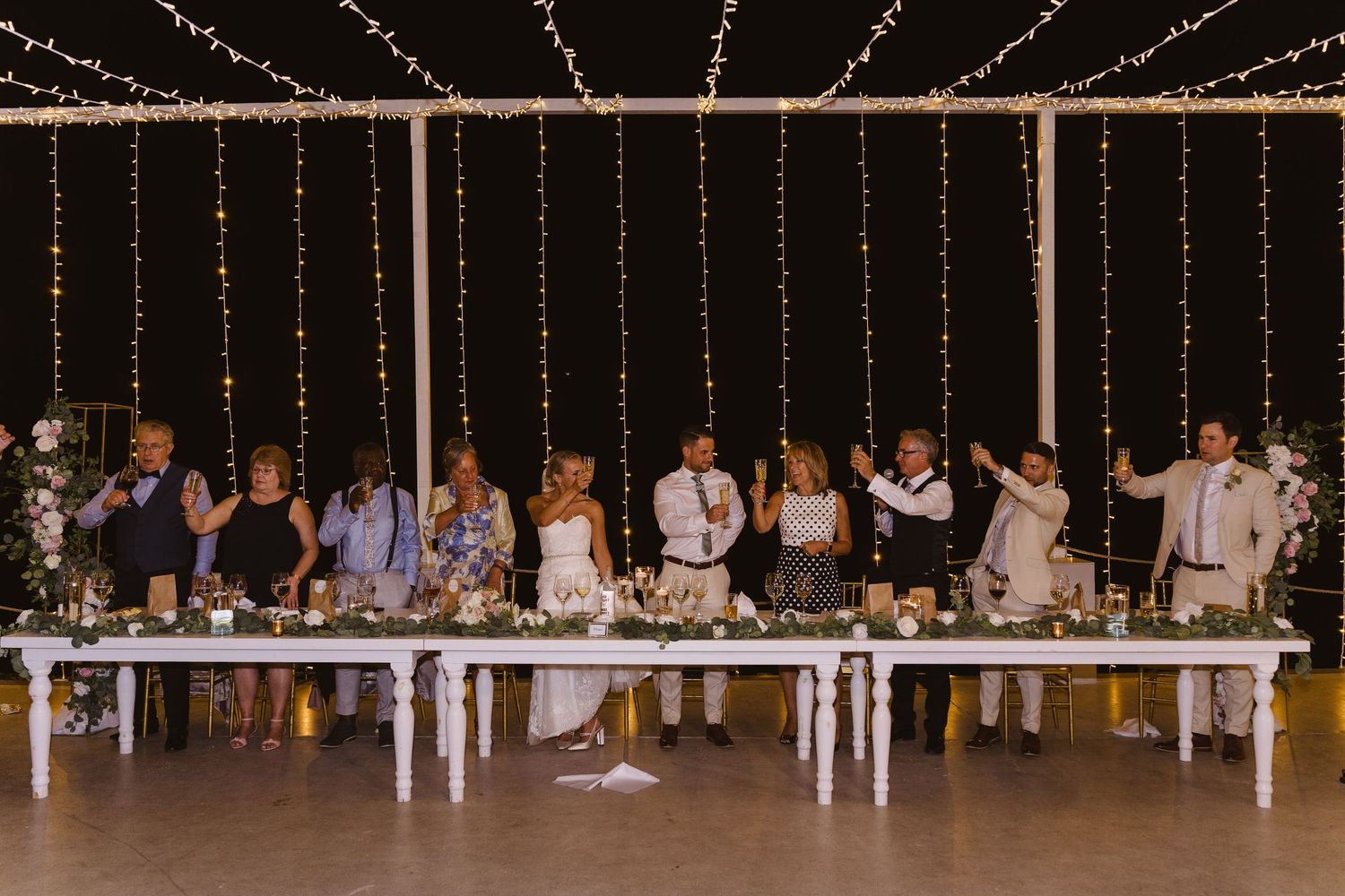 Wedding party seated at long white tables under romantic string lights during evening reception.