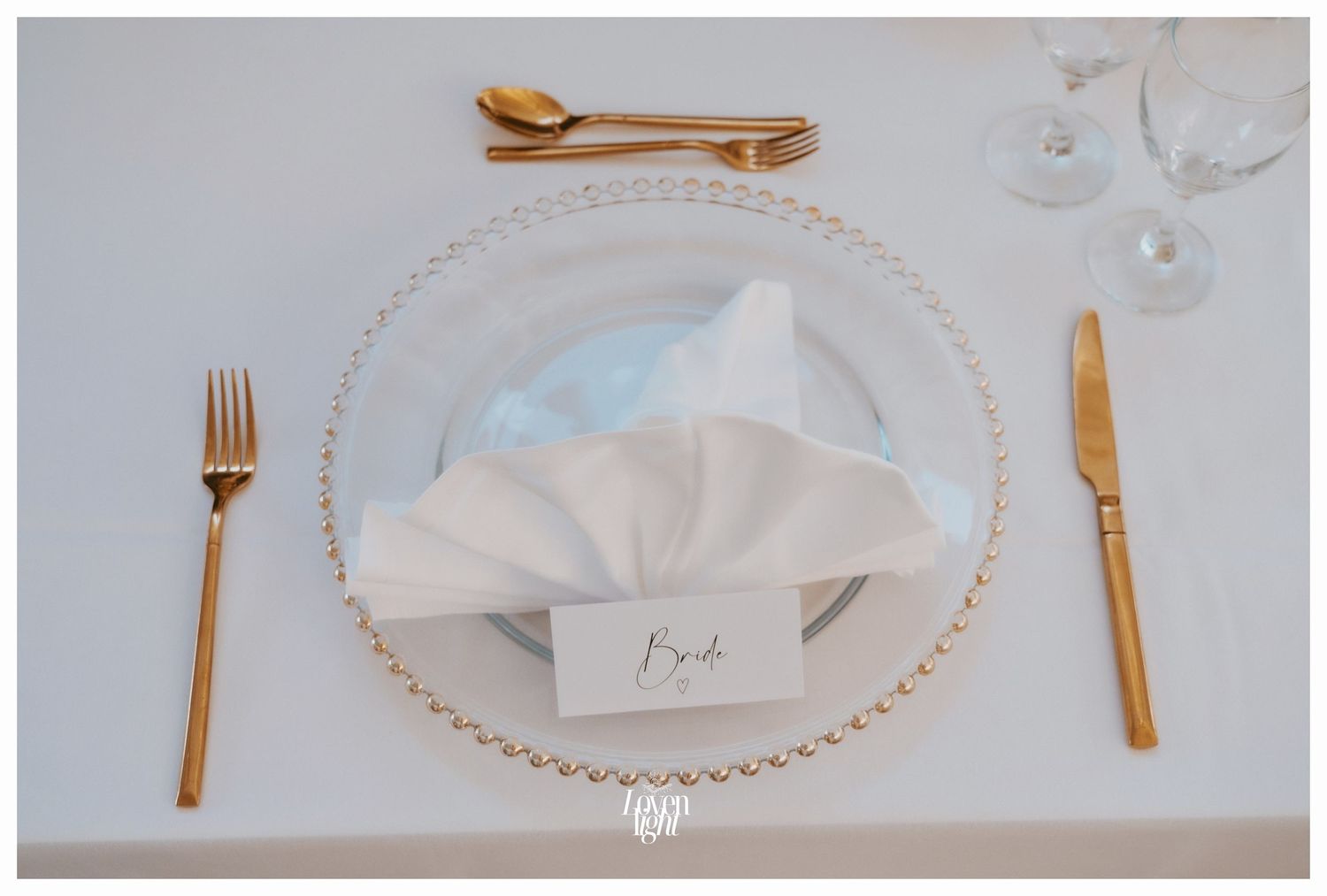 Minimalist wedding table setting with gold utensils and white napkin on beaded glass charger plate.