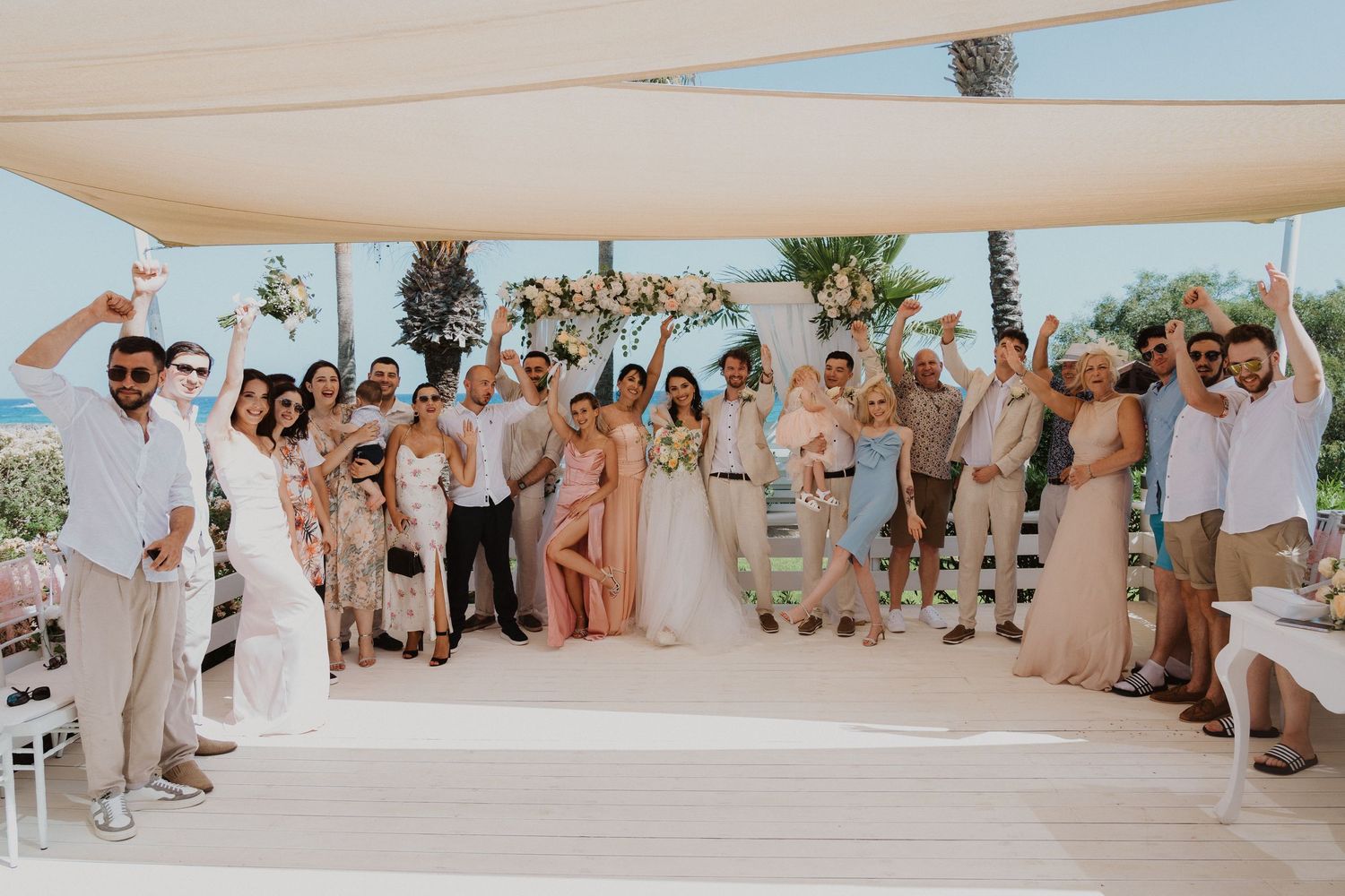 Large wedding group photo with guests in light summer attire gathered under covered oceanfront venue.