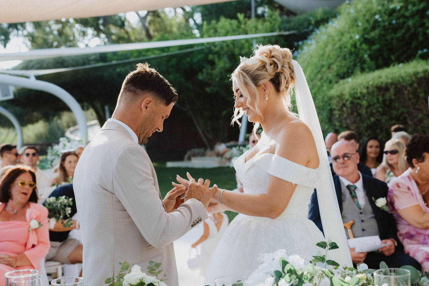 A touching moment during a wedding ceremony as rings are exchanged under an outdoor canopy.