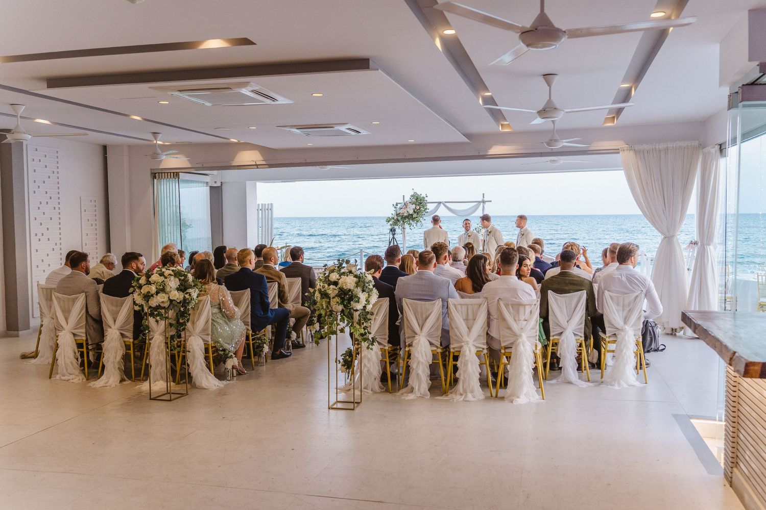 Wedding ceremony setup in a modern venue with ocean views and rows of white chairs for guests.