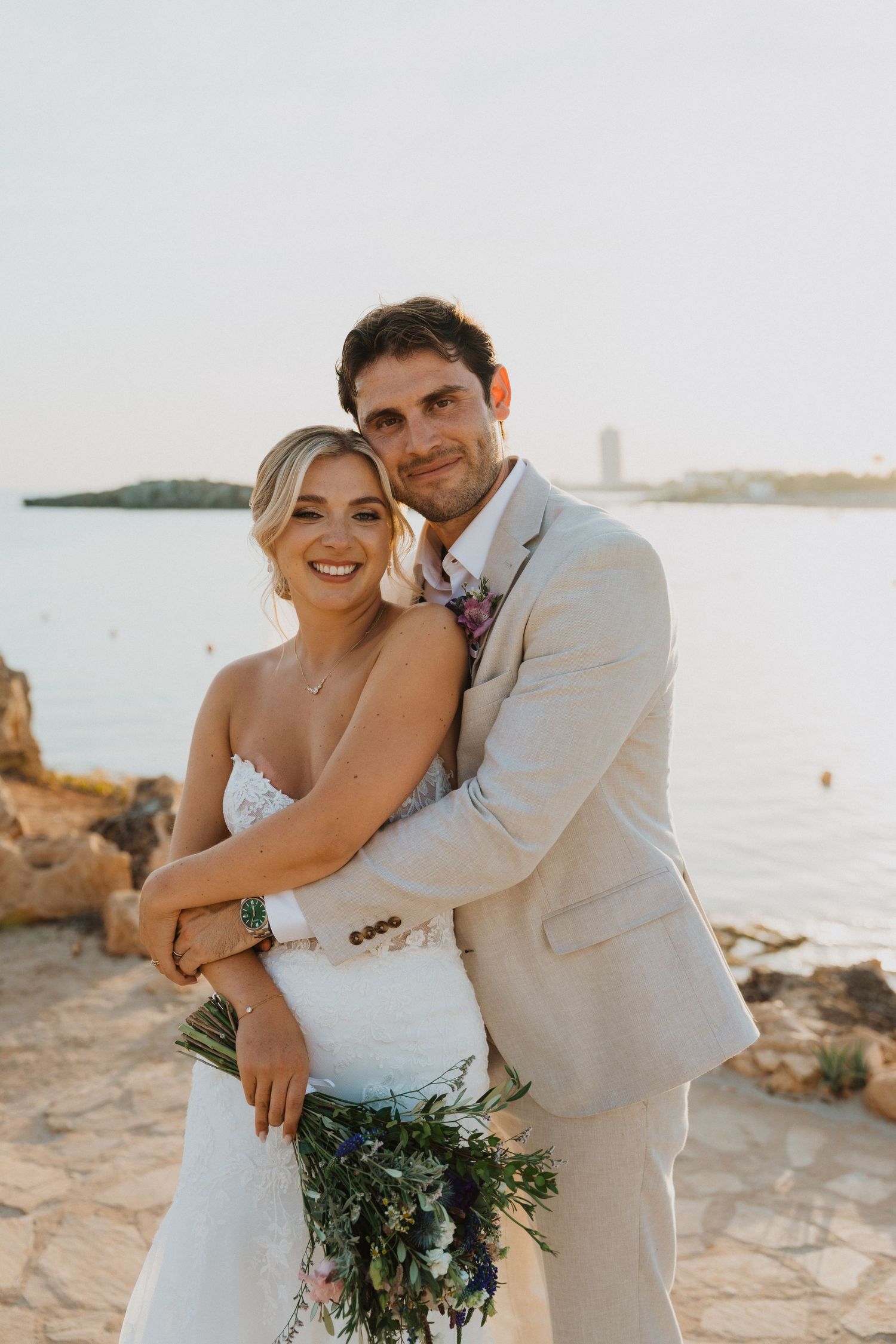 Happy couple embraces on coastal rocks during sunset wedding photoshoot.