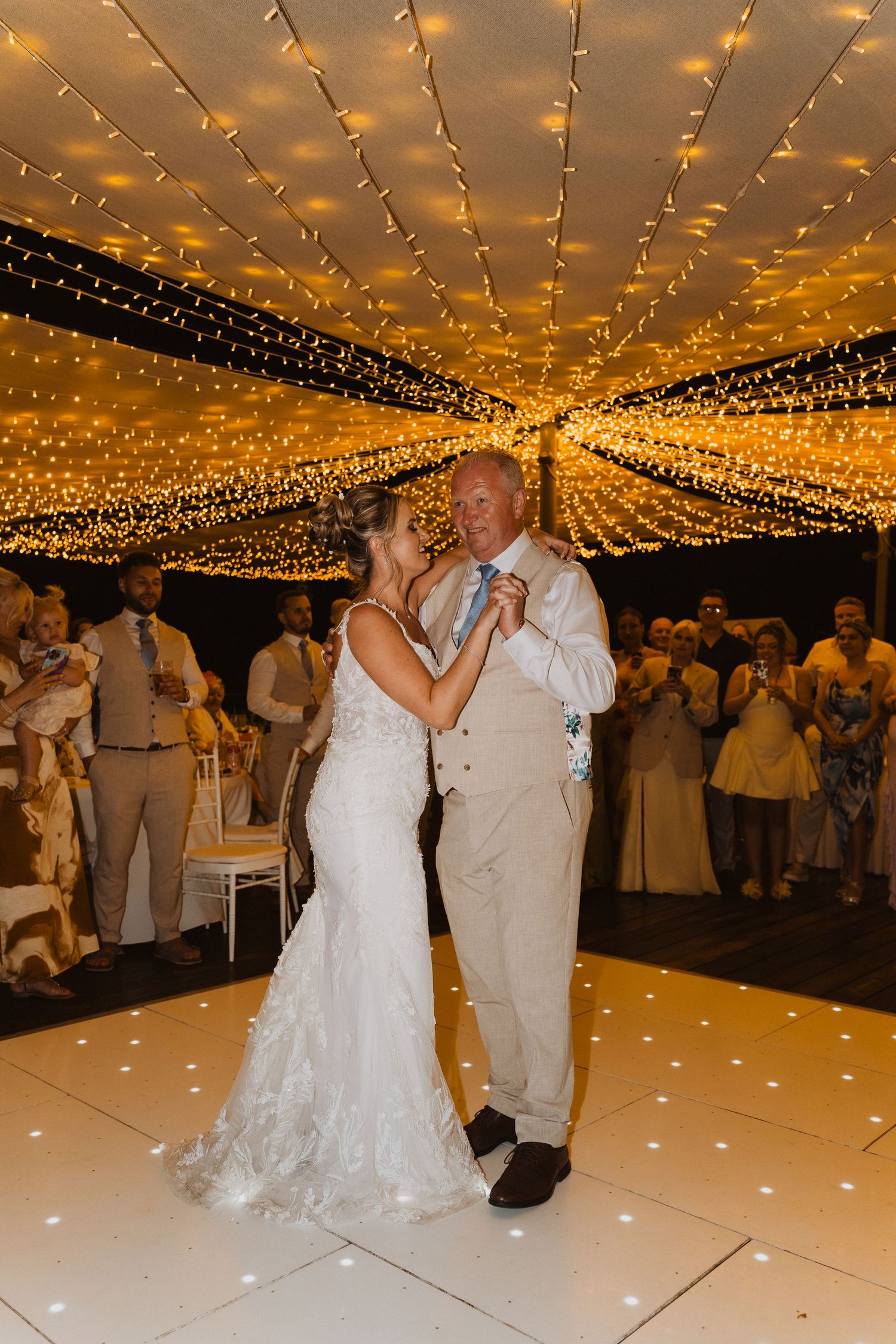 A couple shares their first dance on an illuminated white dance floor under romantic twinkle lights at their wedding reception.