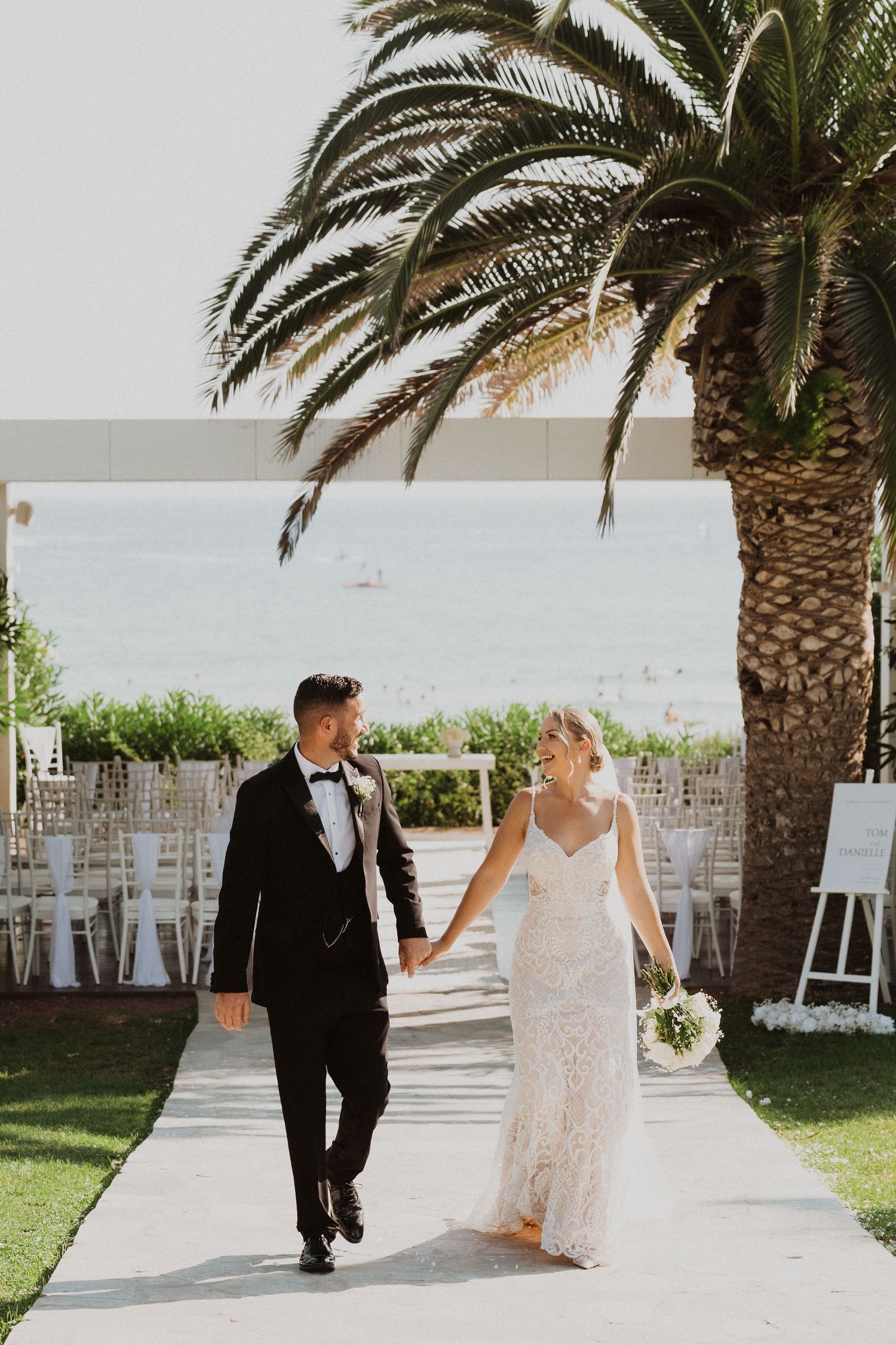 Wedding couple walks along palm-lined pathway at outdoor venue.