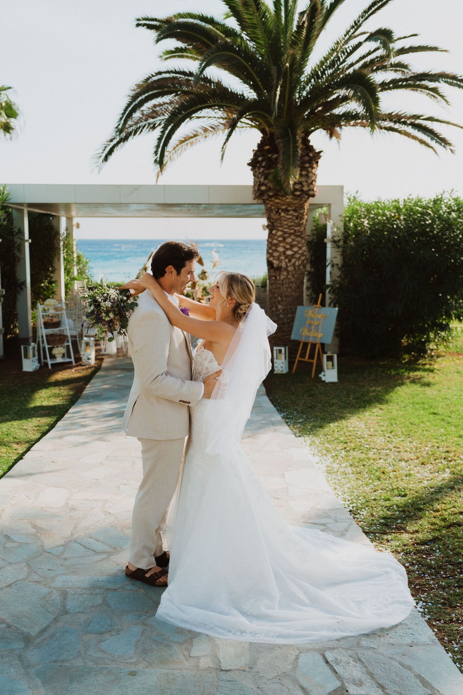 A tender wedding moment captured on a stone pathway with a palm tree and ocean view.