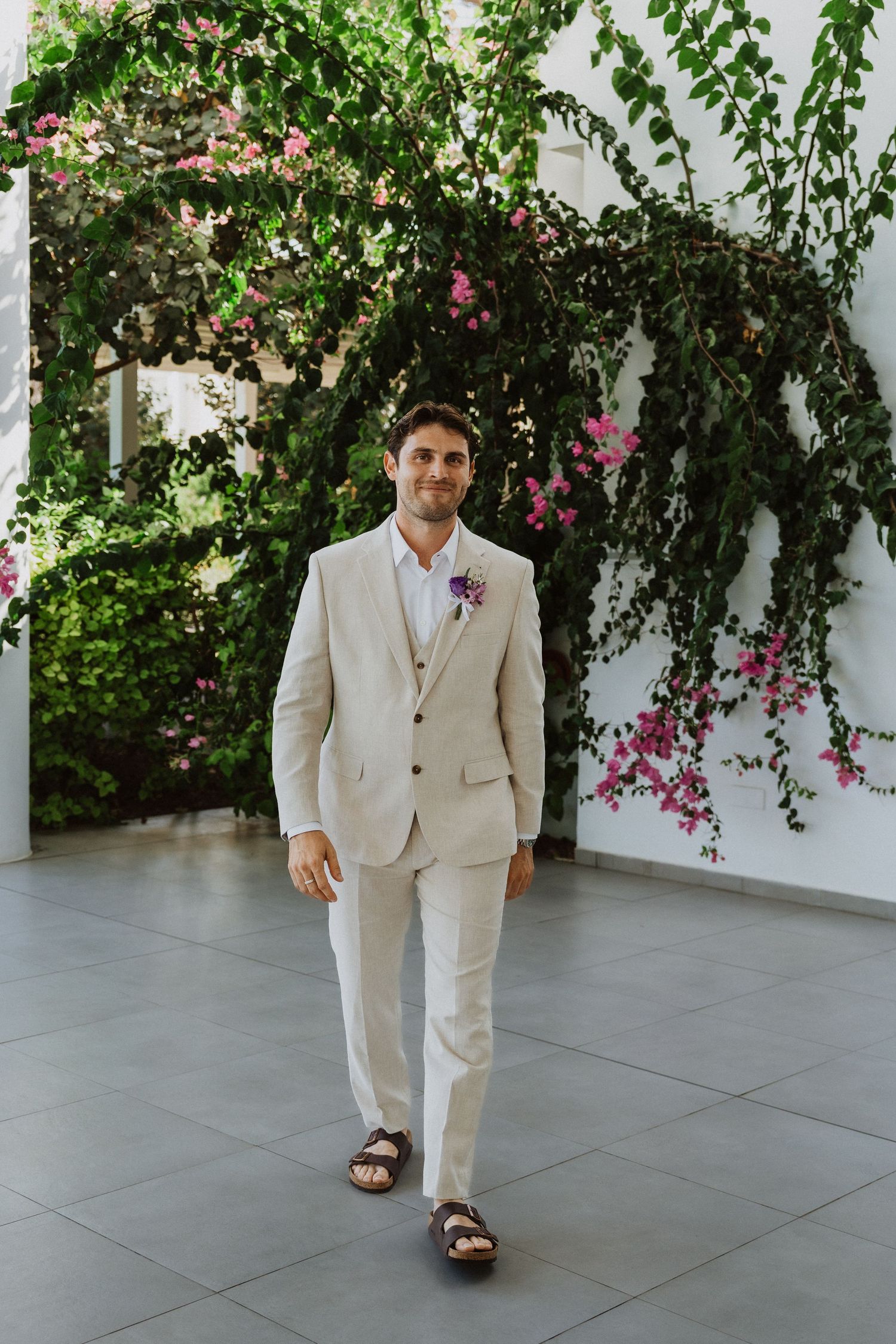 Groom in light beige suit walks along tiled pathway with flowering vines on wall.