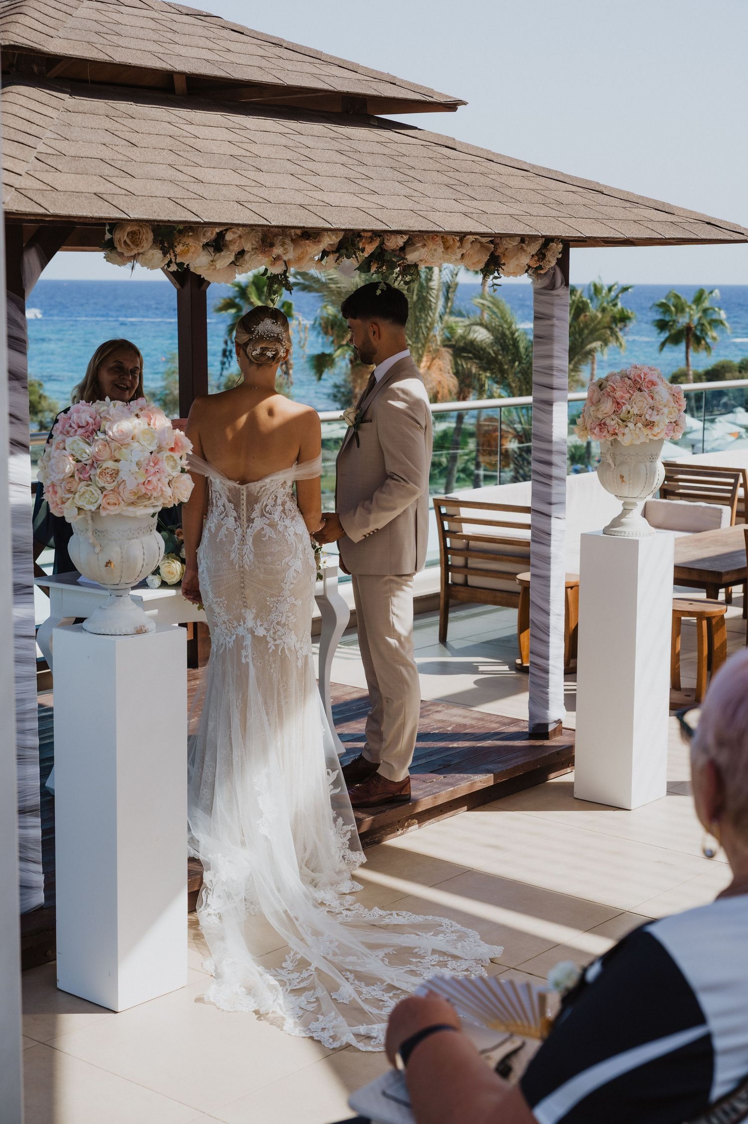An elegant beach wedding setup features white columns with floral arrangements and ocean views.