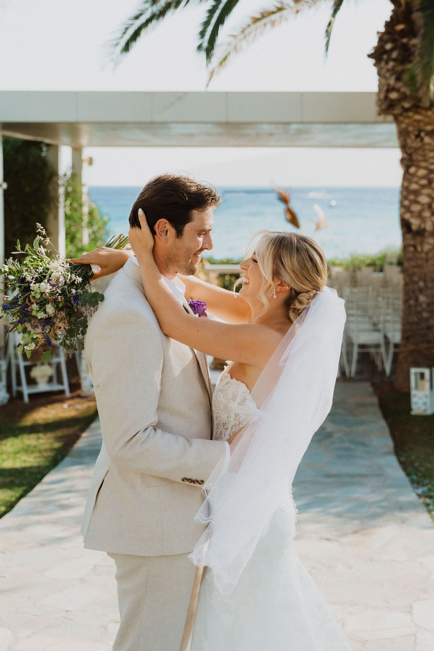 Romantic wedding couple embraces during their beachfront ceremony with palm trees and ocean views.