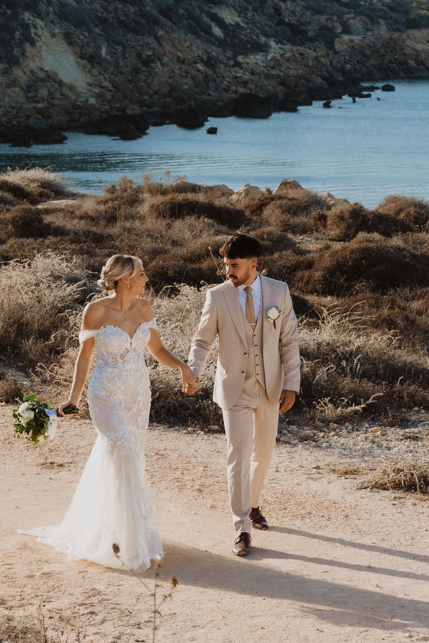 A couple walks along a scenic coastal path during their wedding photoshoot with rocky cliffs and ocean views.