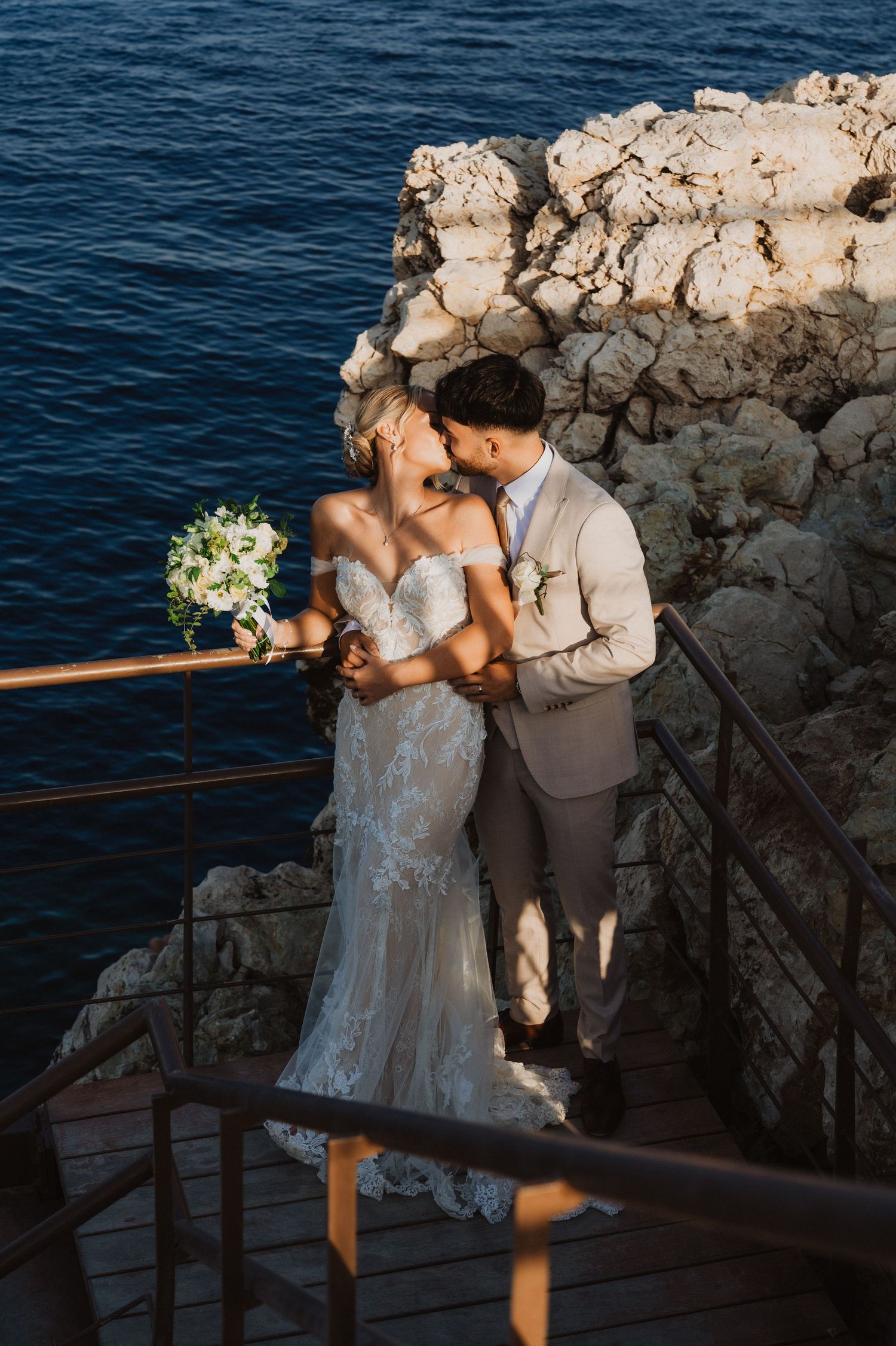 Newlyweds share tender moment on coastal boardwalk with rocky cliffs and deep blue sea.