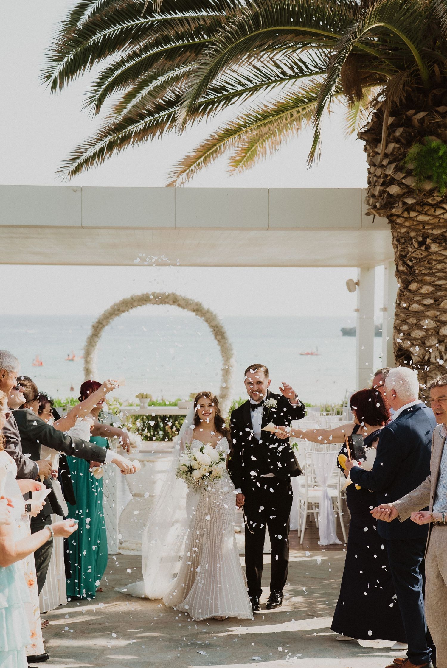 Joyful newlyweds walk through confetti shower after outdoor beach wedding ceremony under floral arch.