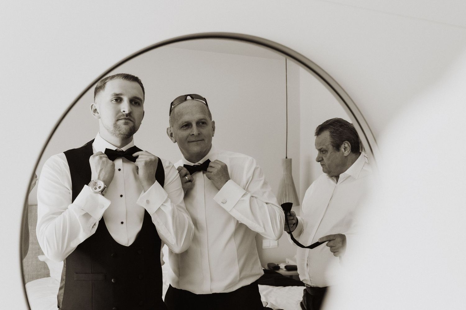Black and white photo of groom adjusting bow tie with groomsman reflected in round mirror.