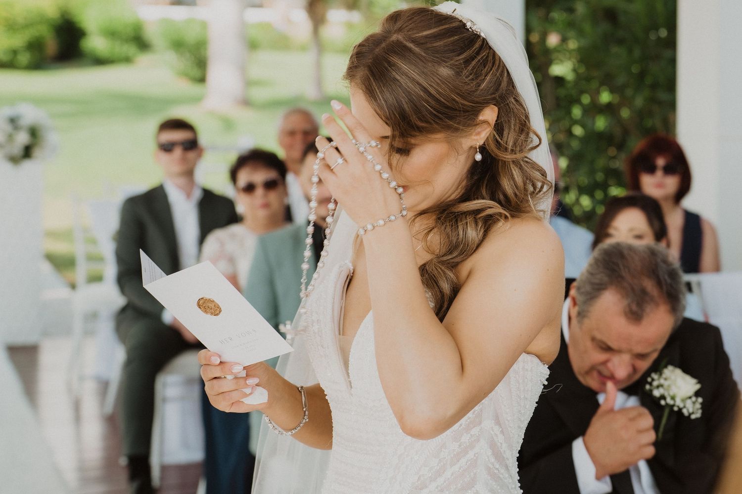 Emotional bride crying while reading wedding vows during outdoor ceremony with guests.