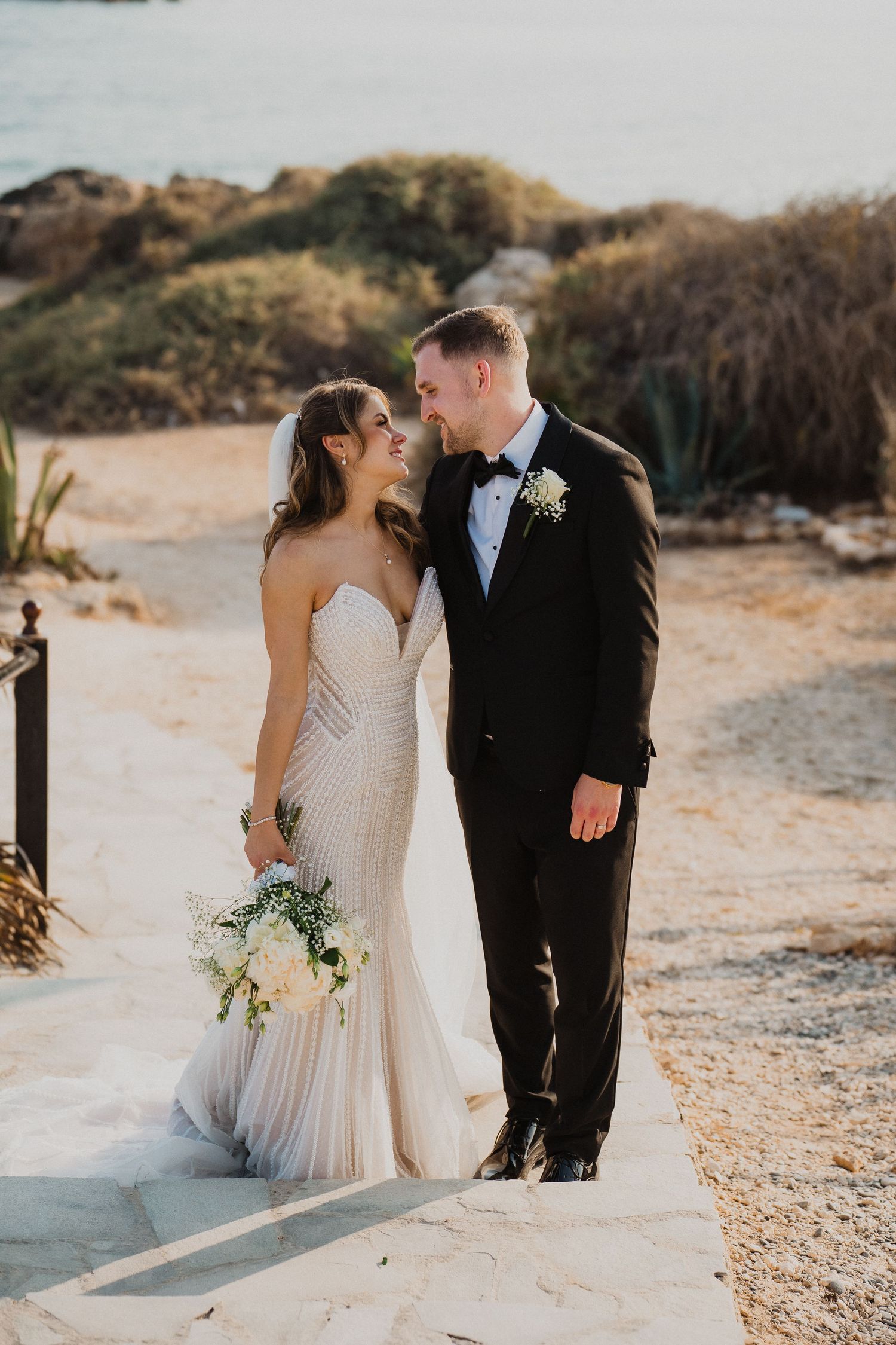 Smiling bride and groom share tender moment on coastal pathway with ocean backdrop at golden hour.